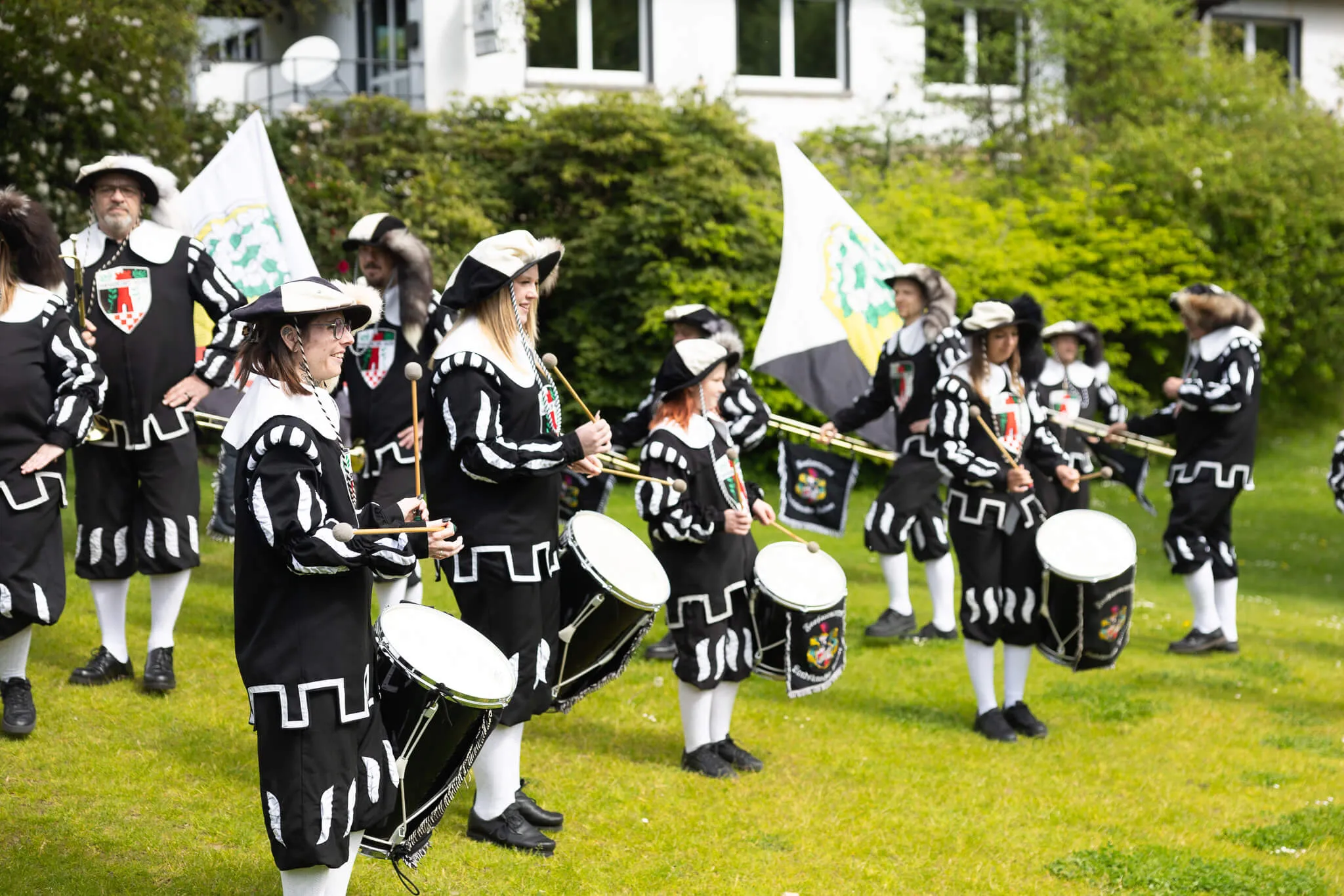 Eine Spielmannszug-Gruppe in historischen Uniformen mit Dreispitz und verzierten Jacken spielt auf Trommeln und Hörnern auf einer grünen Wiese.