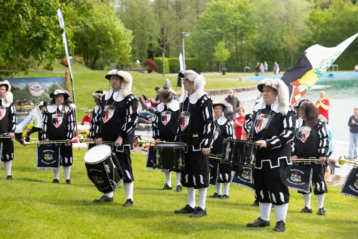 Mehrere Musiker in historischen Uniformen mit Trommeln und Fahnen stehen auf einer grünen Wiese vor Zuschauern.