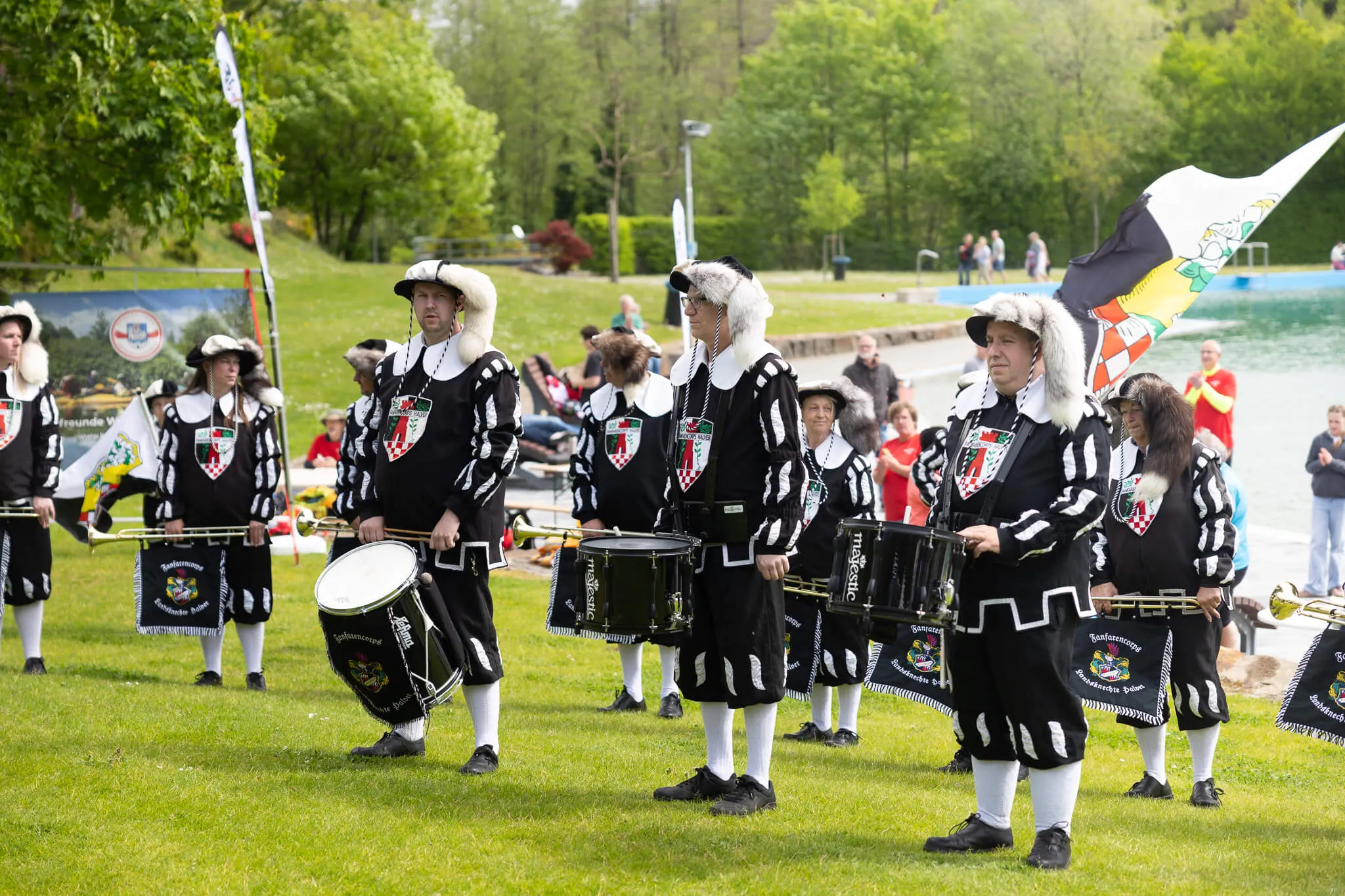 Mehrere Musiker in historischen Uniformen mit Trommeln und Fahnen stehen auf einer grünen Wiese vor Zuschauern.