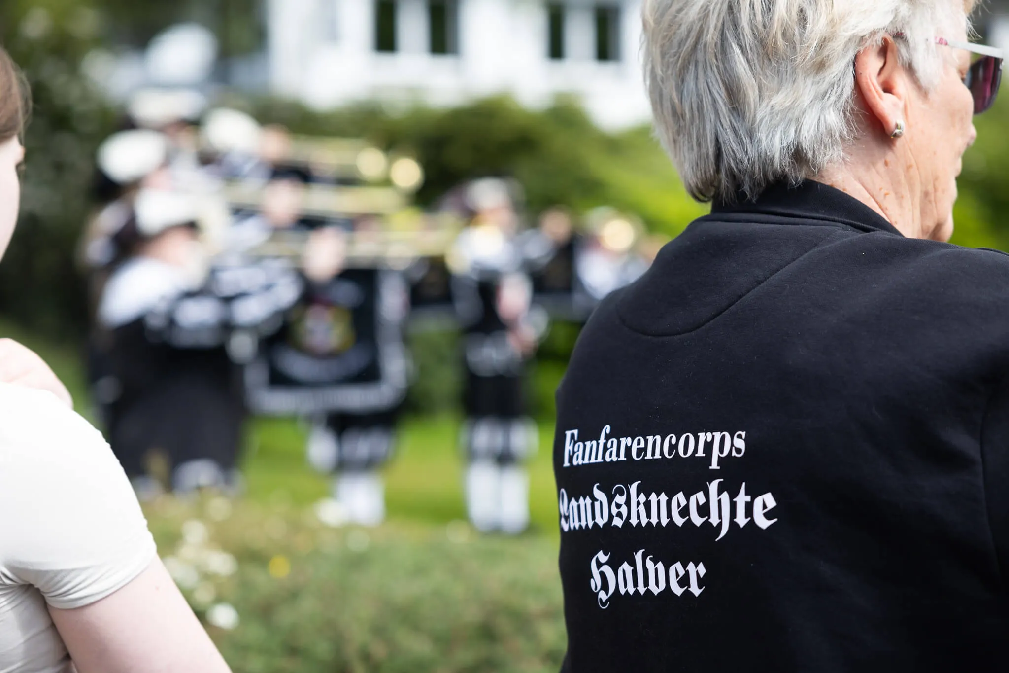Rückansicht einer Frau mit kurzem, grauem Haar, die auf eine Gruppe in historischen Uniformen blickt. Auf ihrem schwarzen Oberteil steht in geschwungener Schrift „Fanfarencorps Hundsknechte Halver“.