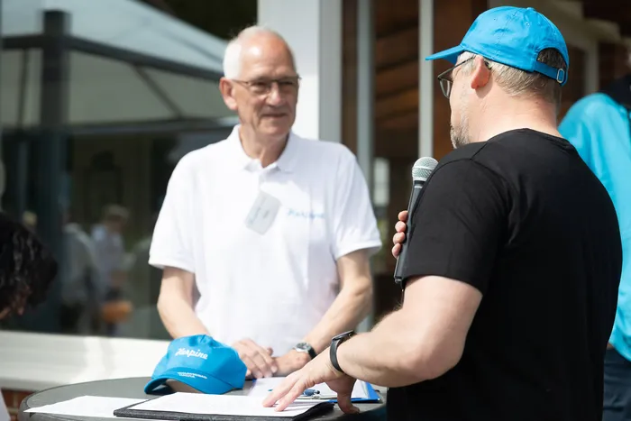 Ein Mann mit blauem Cap hält ein Mikrofon und spricht einen älteren Mann in weißem Polohemd an. Im Vordergrund liegt eine blaue Cap mit dem Schriftzug „Herpine“ und Notizen auf einem Tisch.