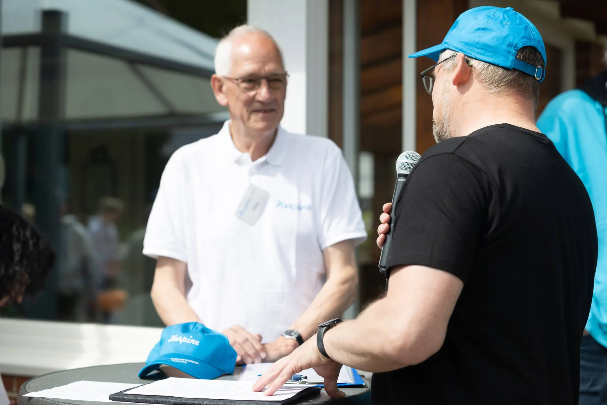 Ein Mann mit blauem Cap hält ein Mikrofon und spricht einen älteren Mann in weißem Polohemd an. Im Vordergrund liegt eine blaue Cap mit dem Schriftzug „Herpine“ und Notizen auf einem Tisch.