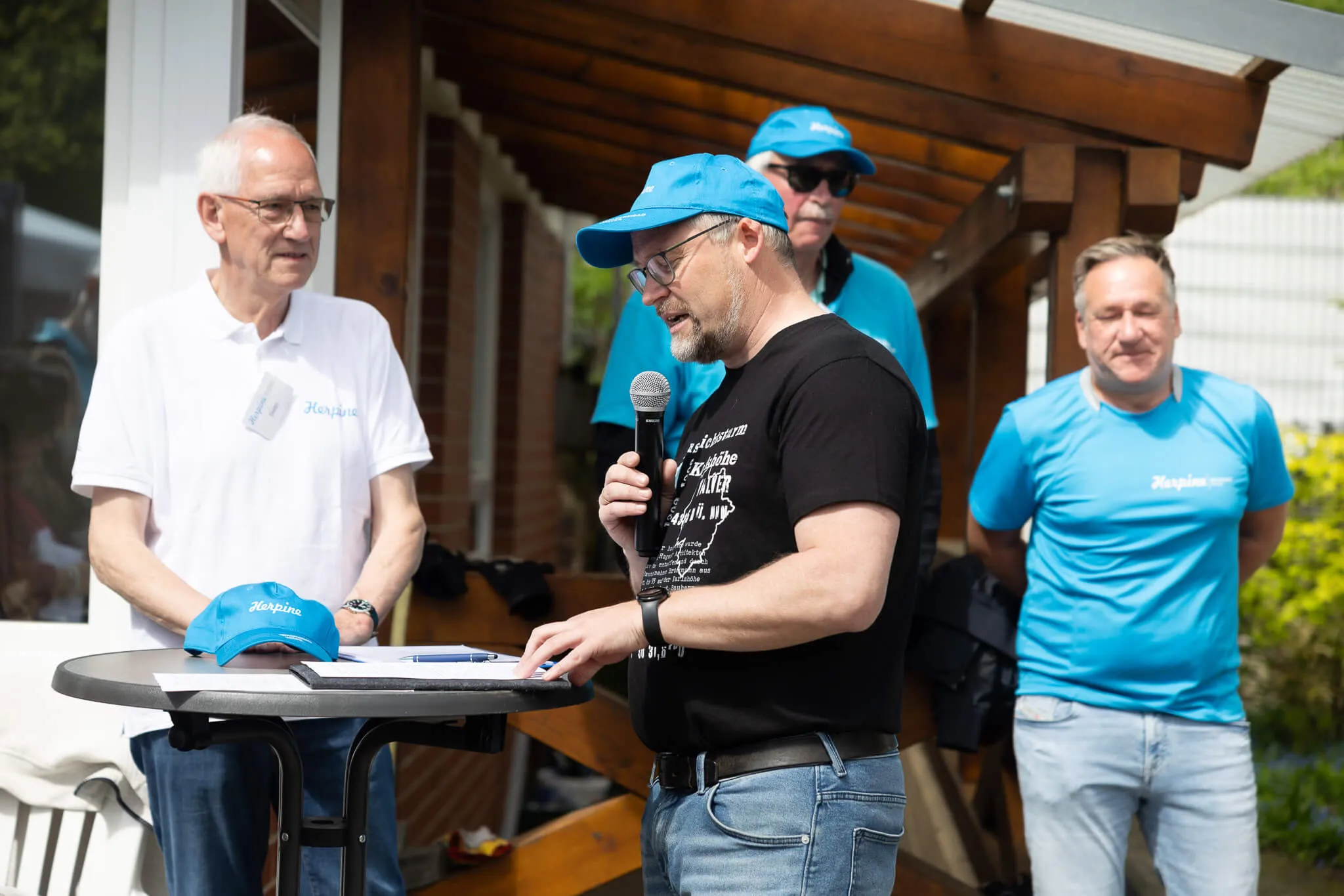 Ein Mann mit blauer Kappe spricht in ein Mikrofon, während er an einem Tisch mit einem Laptop steht. Im Hintergrund stehen weitere Personen, einige tragen T-Shirts mit dem Aufdruck „Herpine“.