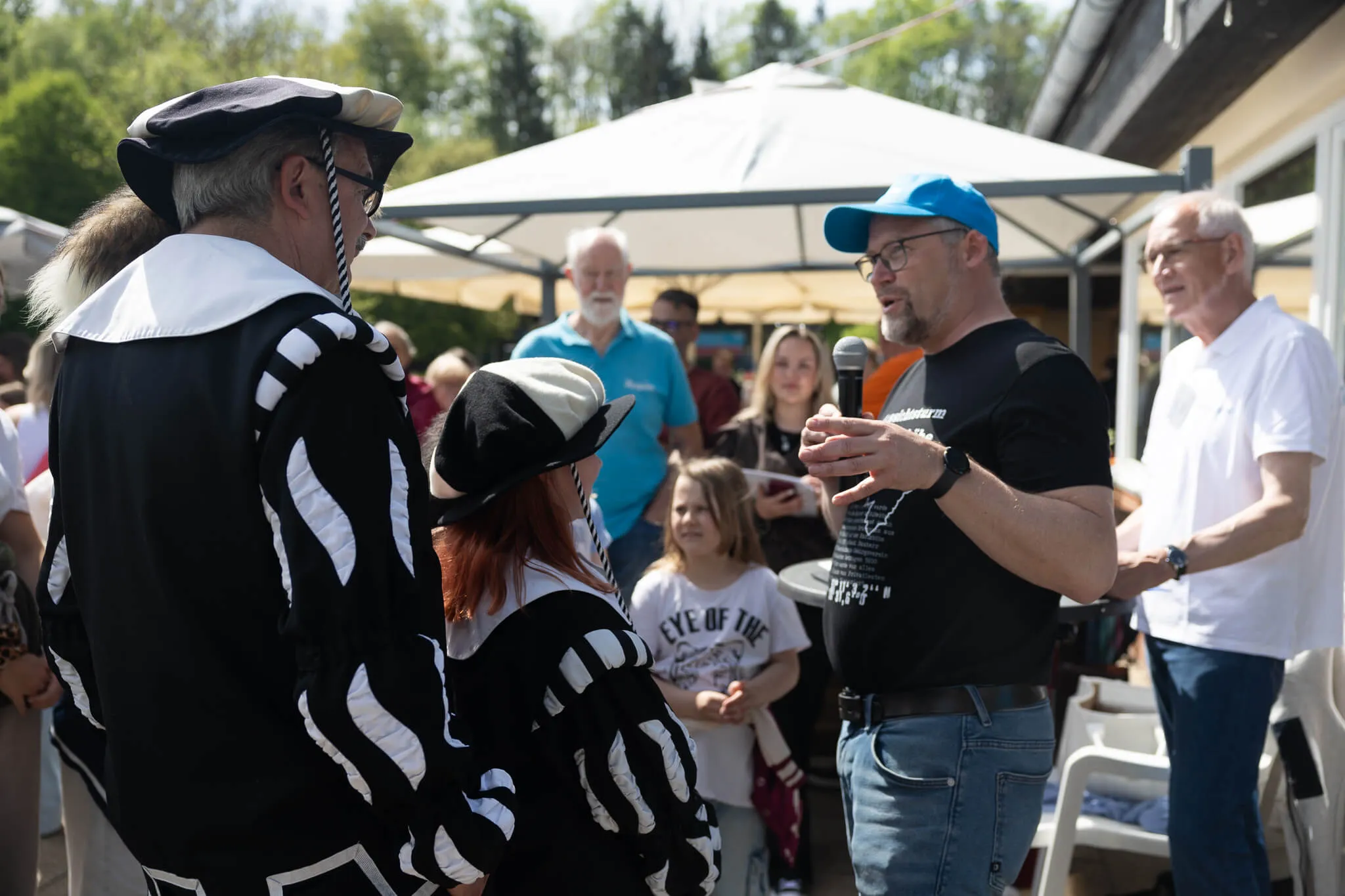 Ein Mann mit blauem Cap spricht in ein Mikrofon, während er sich zu einer Frau im Piratenkostüm umdreht. Im Hintergrund stehen weitere Menschen auf einem sonnigen Platz.