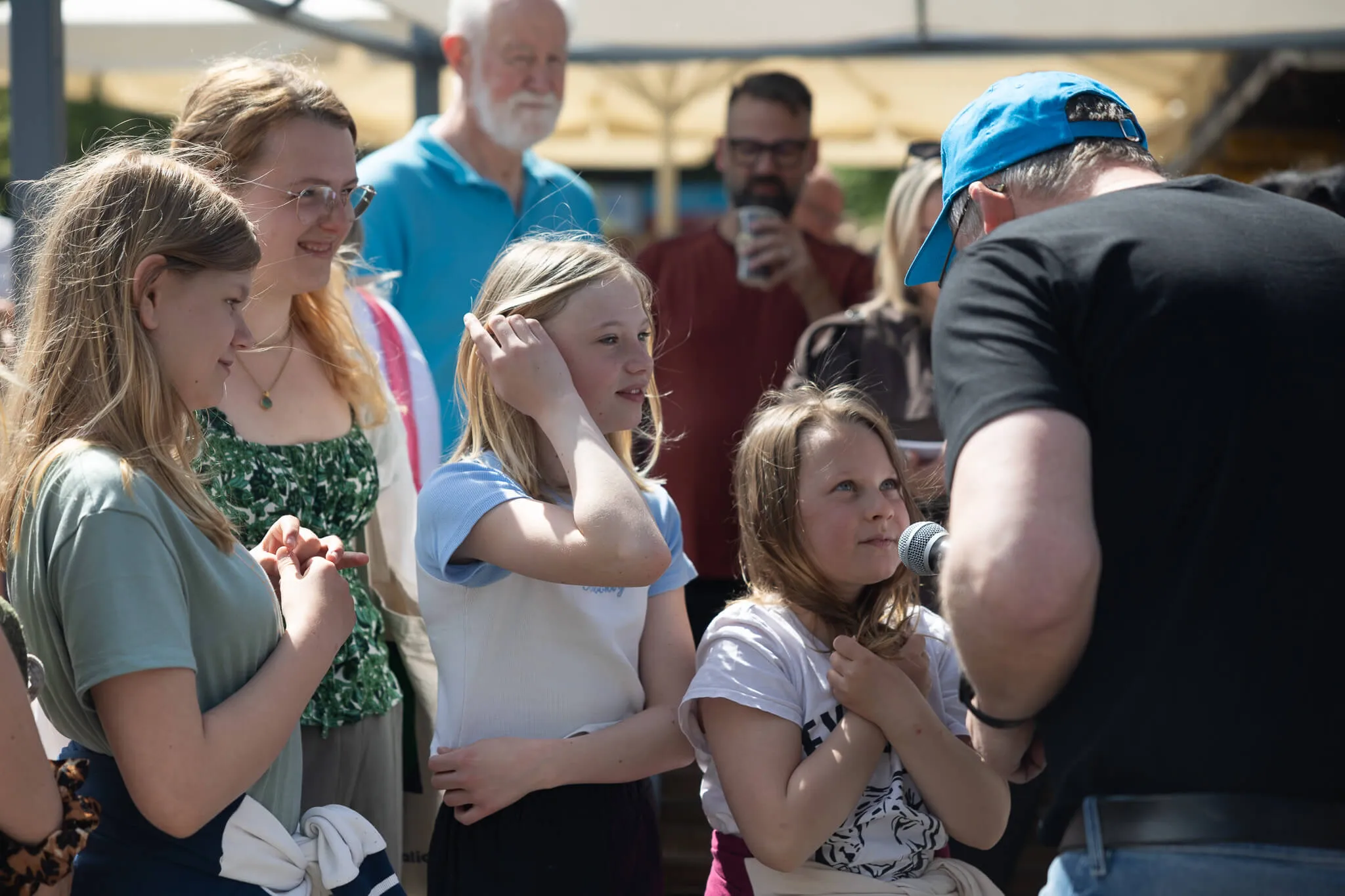 Mehrere junge Frauen und Mädchen stehen vor einem Mann mit blauer Kappe, eine der Mädchen hält ein Mikrofon. Im Hintergrund stehen weitere Personen, darunter ein älterer Mann mit Bart.