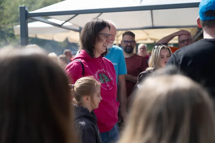 Eine Frau mit roten Haaren lächelt in die Kamera, während sie auf einem Freiluftfest steht. Im Hintergrund sind weitere Menschen und ein weißes Zelt zu sehen.