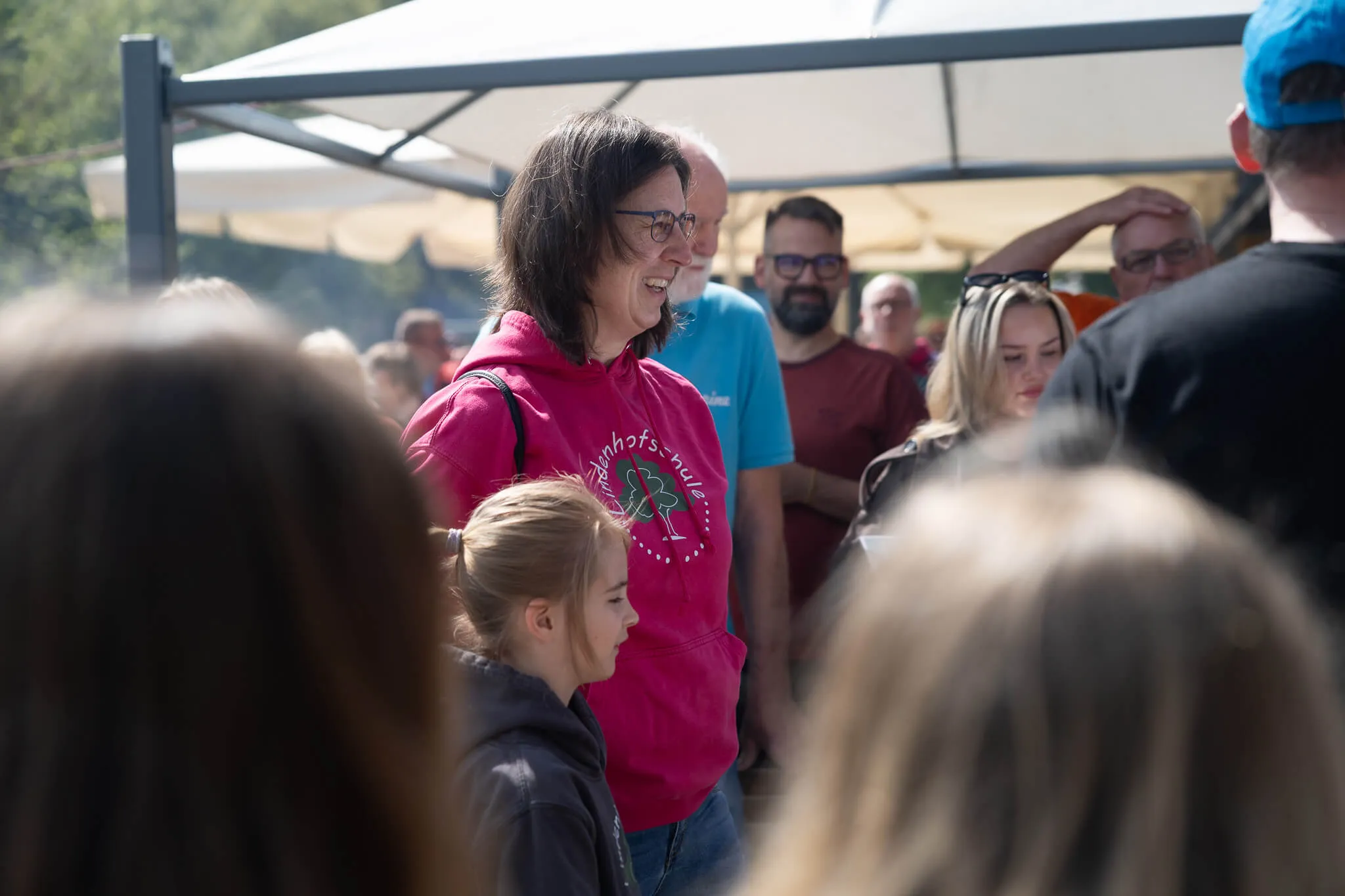 Eine Frau mit roten Haaren lächelt in die Kamera, während sie auf einem Freiluftfest steht. Im Hintergrund sind weitere Menschen und ein weißes Zelt zu sehen.