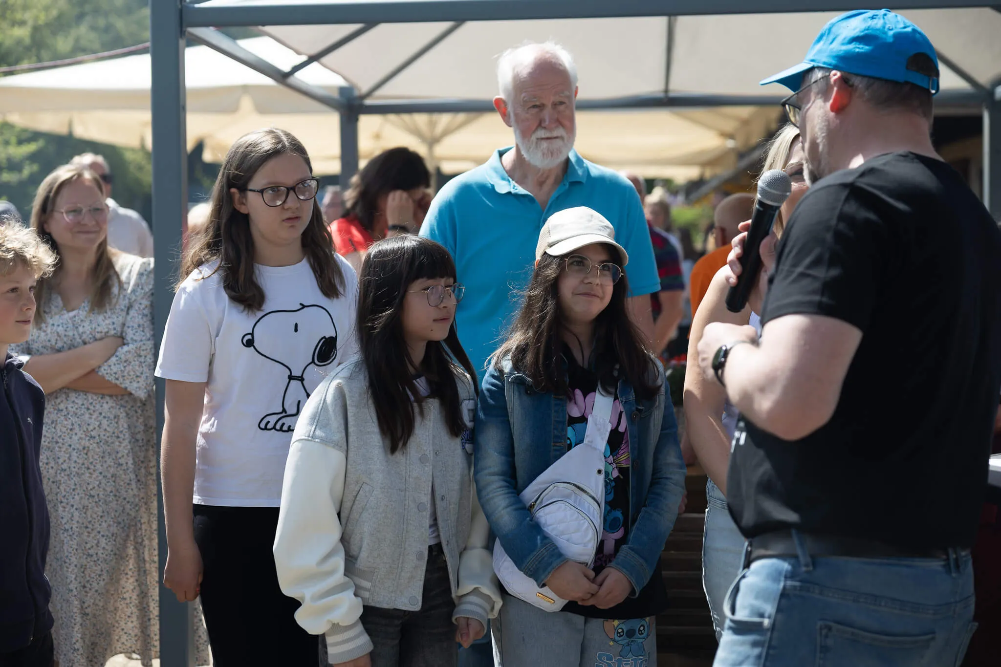 Drei Mädchen unterschiedlichen Alters stehen vor einem Mann mit Mikrofon, im Hintergrund weitere Menschen und ein Pavillon.