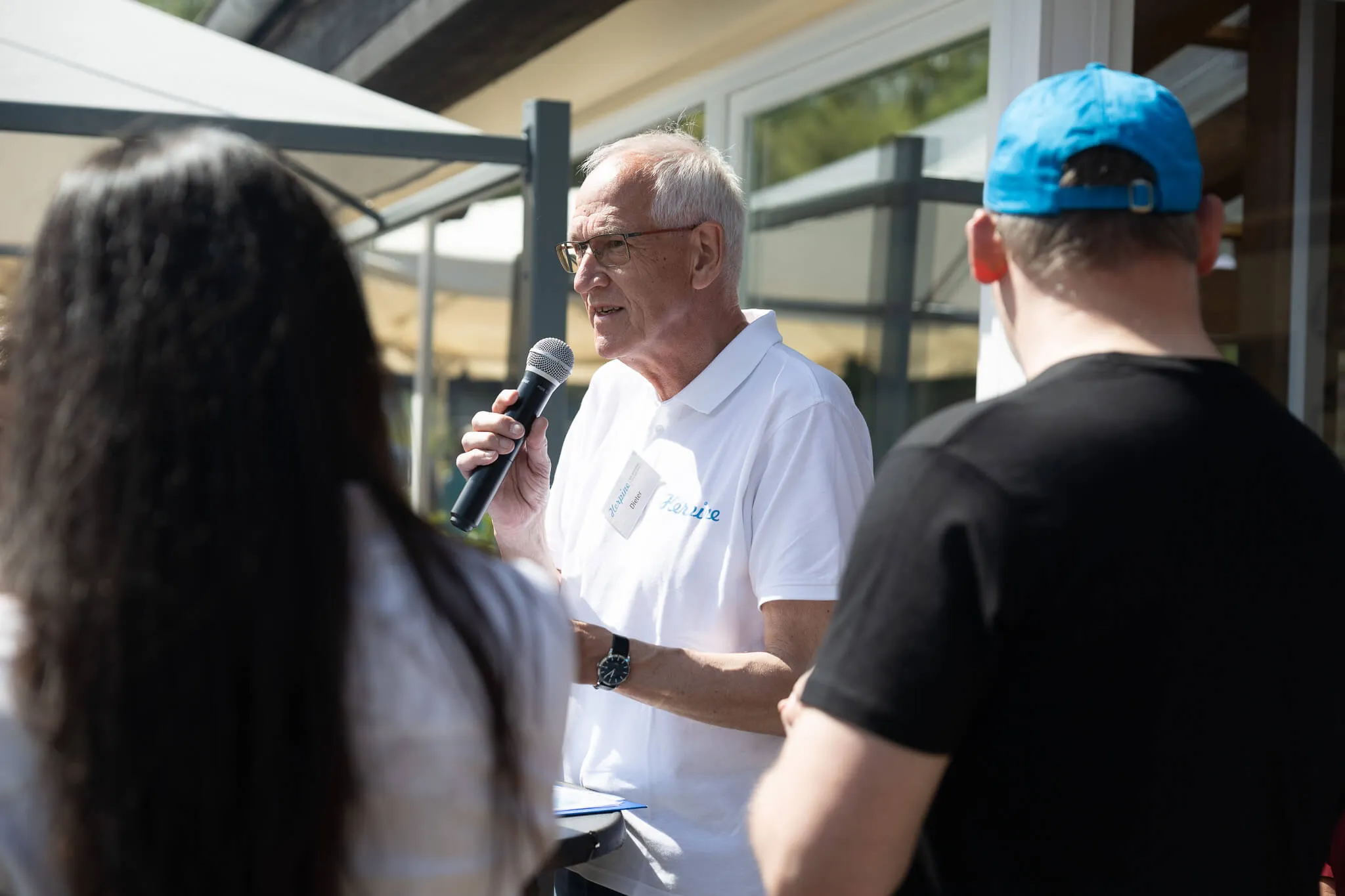 Ein älterer Mann mit weißem Haar hält ein Mikrofon und spricht vor einer Gruppe von Menschen im Freien. Er trägt ein weißes Polohemd mit dem Schriftzug „Herpine“ und eine Armbanduhr.