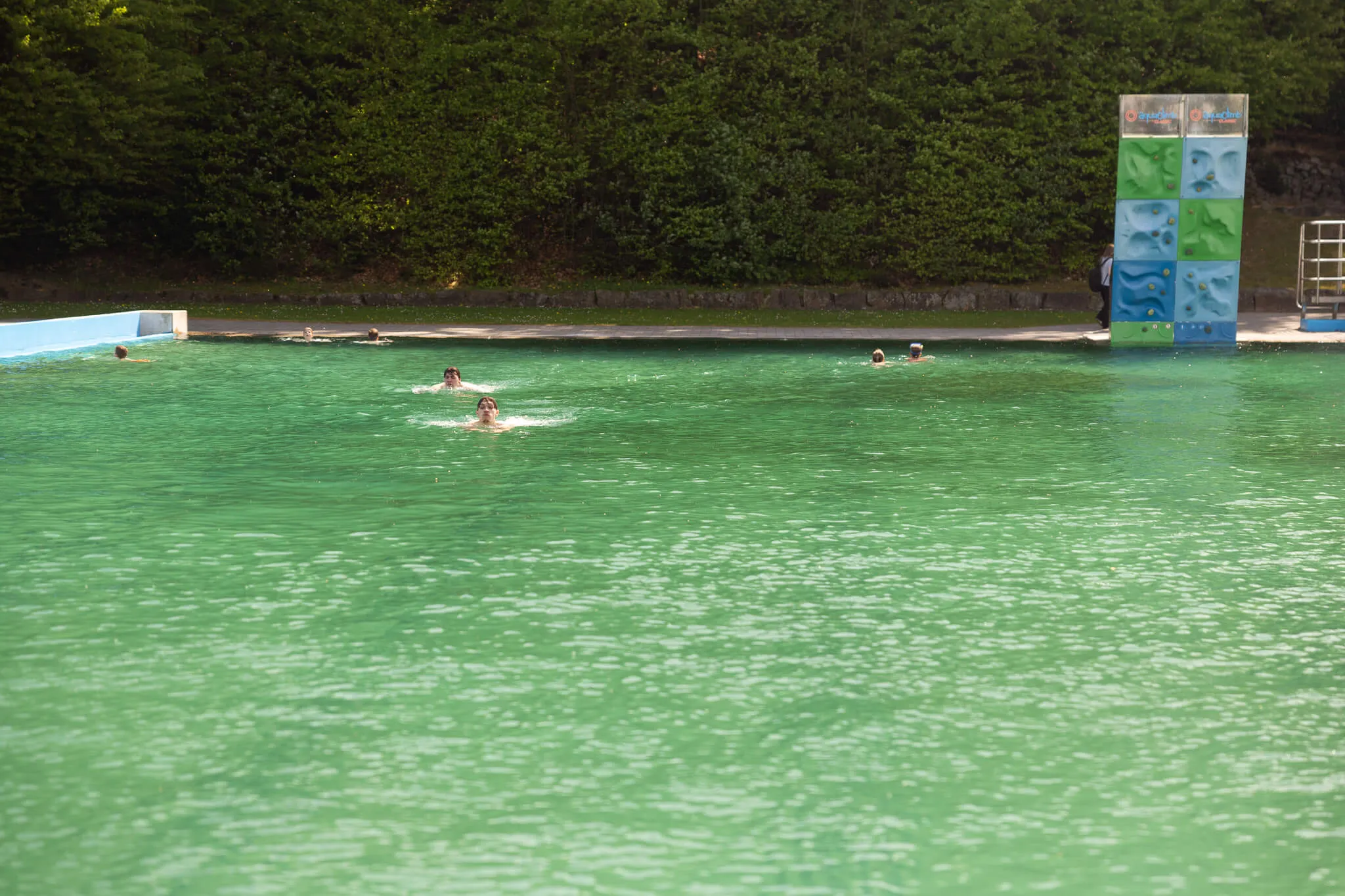 Menschen schwimmen in dem türkisfarbenen Wasser des Freibads Herpine in Halver. Im Hintergrund ein bewaldeter Hang und eine Kletterwand.