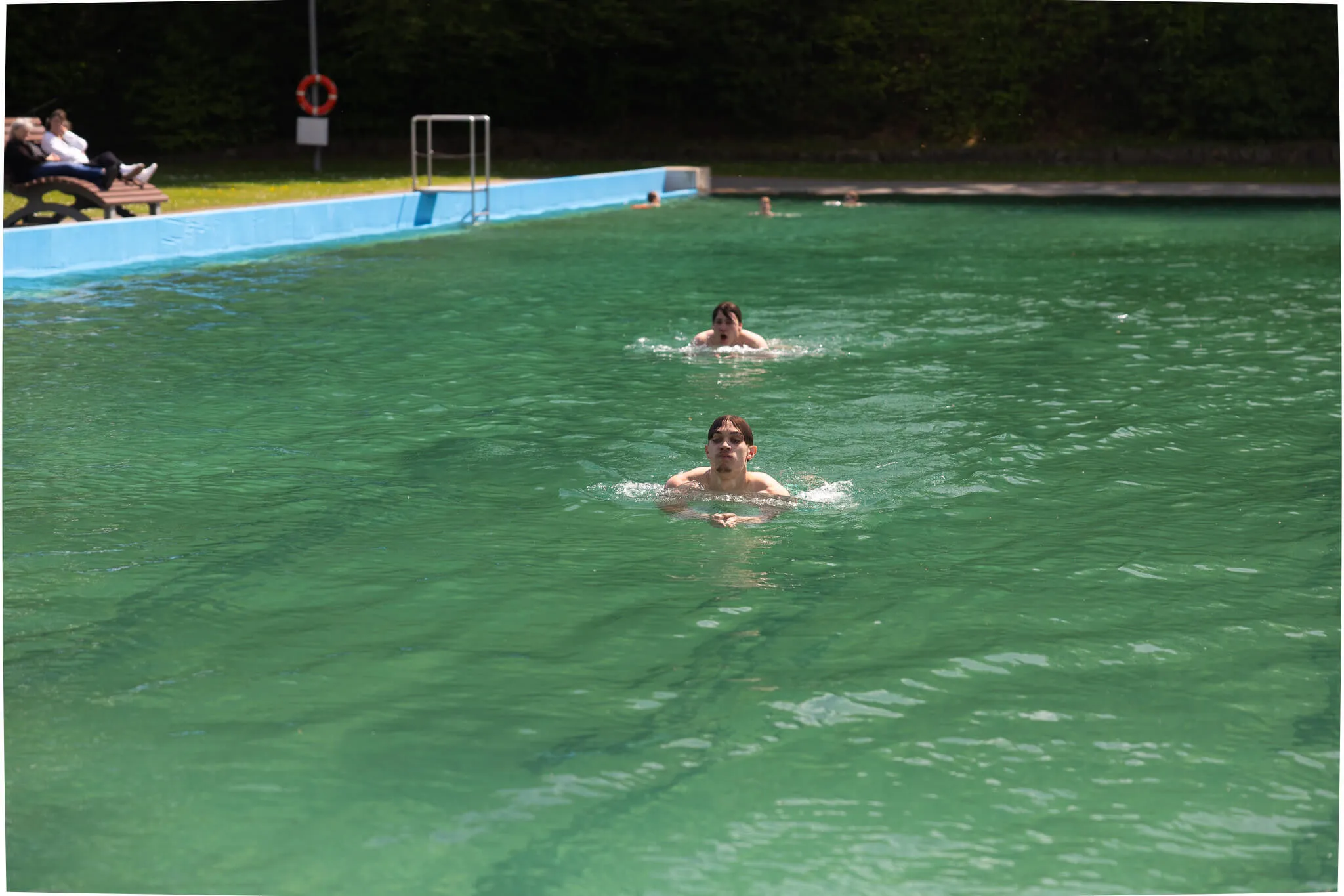 Zwei Schwimmer im türkisfarbenen Wasser der Herpine Halver, im Hintergrund eine weitere Person auf einer Bank.