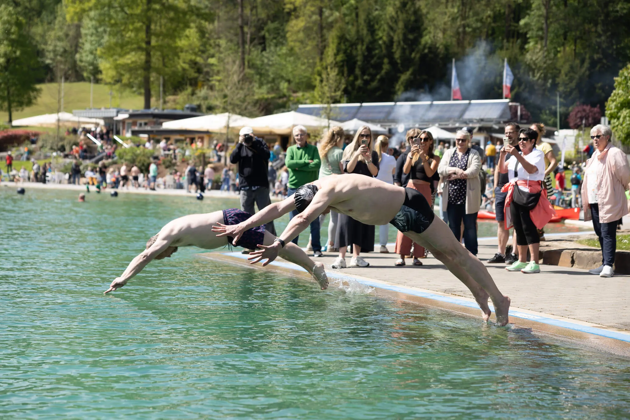 Zwei Männer springen in den See, während Zuschauer am Ufer stehen. Im Hintergrund sind Bäume und Gebäude zu sehen.