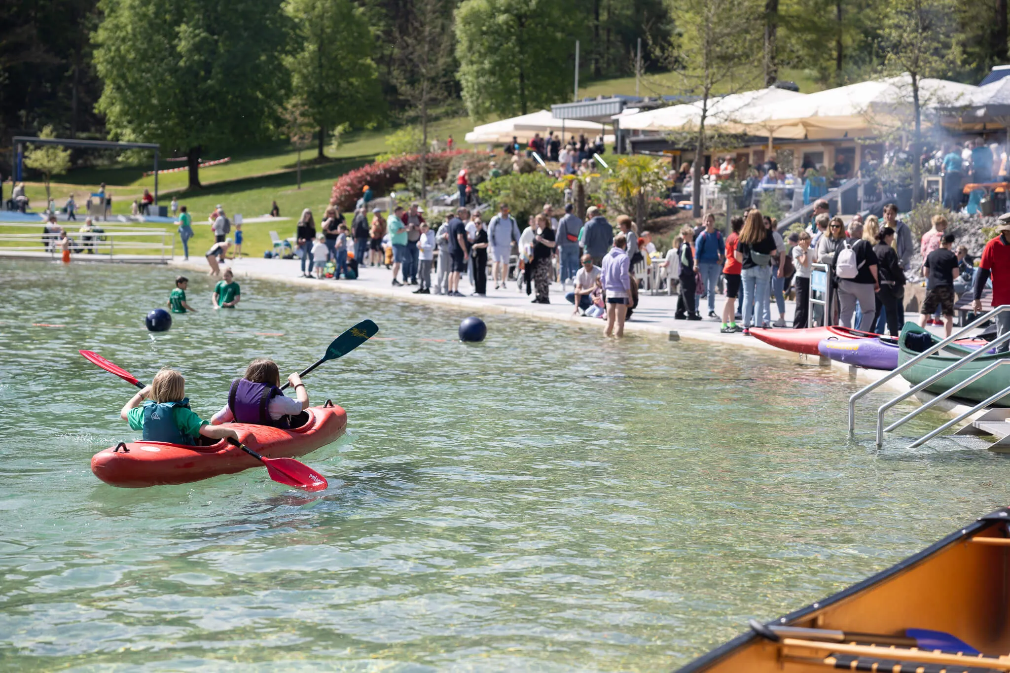 Zwei Personen im roten Kajak paddeln auf dem See, während im Hintergrund Menschen an Land stehen und sich unterhalten. Im Hintergrund ein Restaurant mit Sonnenschirmen.