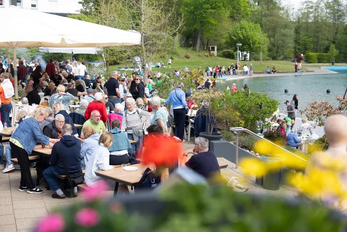 Viele Menschen sitzen an Tischen vor einem Gebäude mit Sonnenschirmen und essen oder unterhalten sich. Im Hintergrund ein See und grüne Bäume.