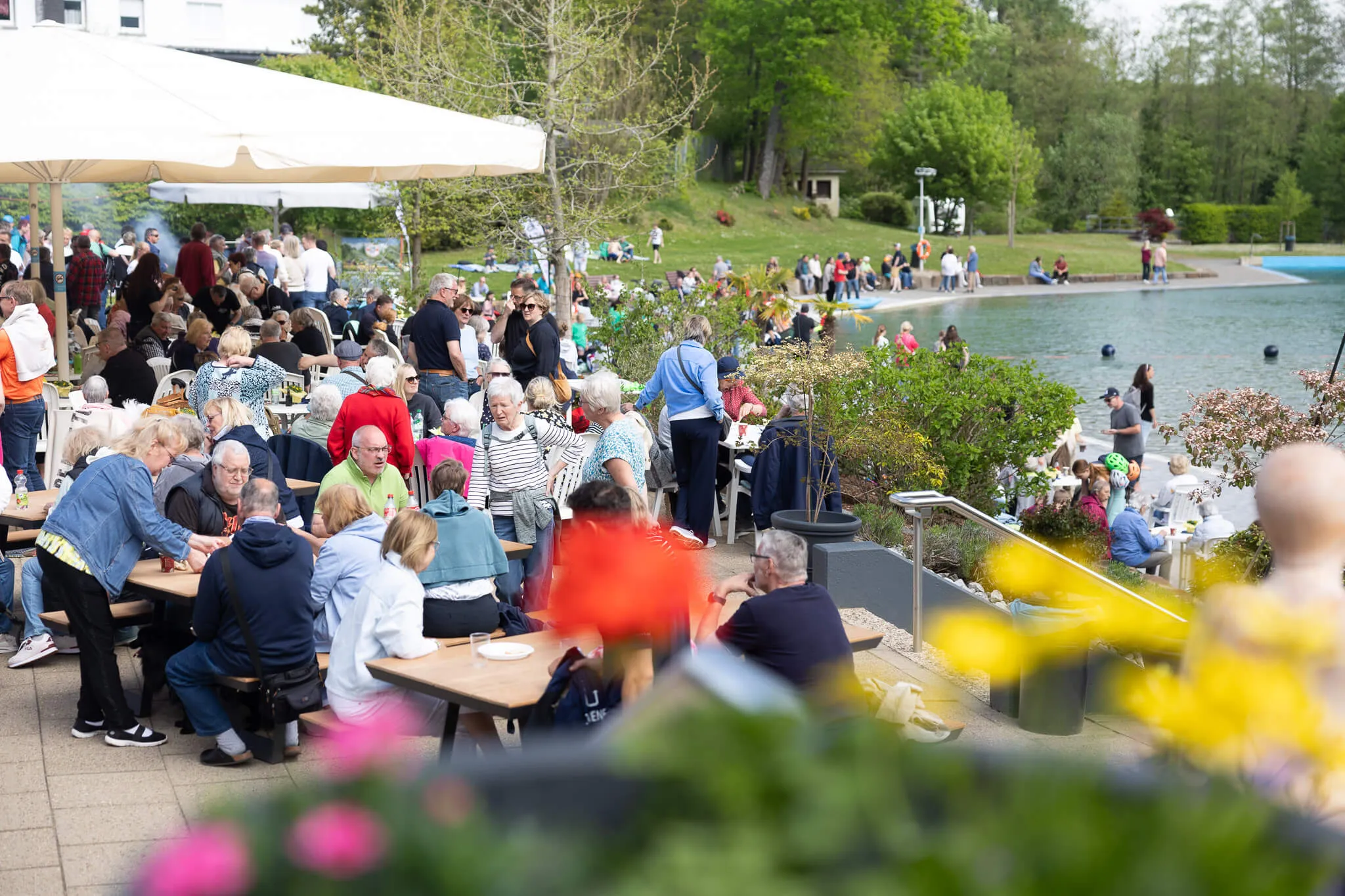 Viele Menschen sitzen an Tischen vor einem Gebäude mit Sonnenschirmen und essen oder unterhalten sich. Im Hintergrund ein See und grüne Bäume.
