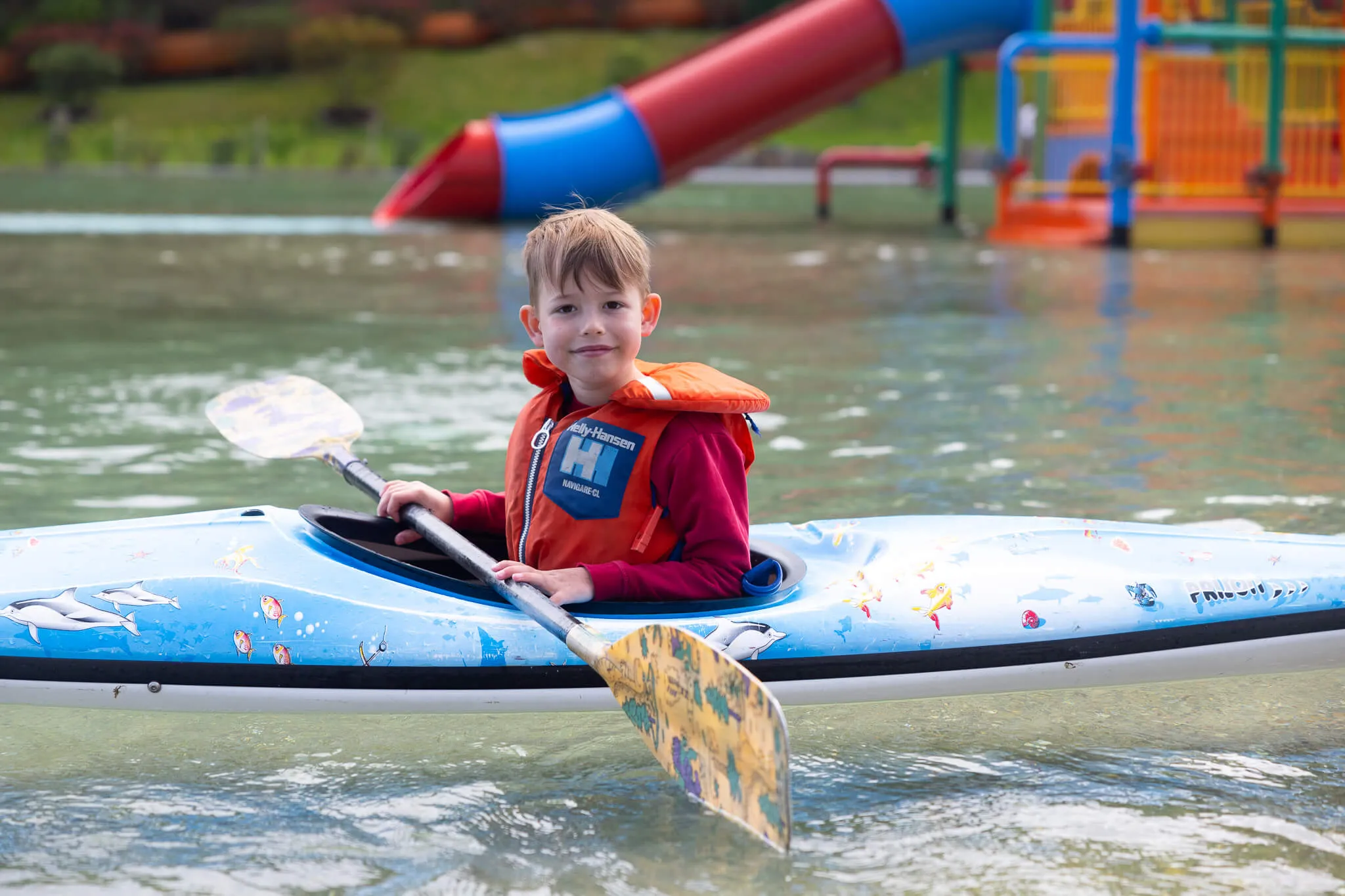 Ein Junge mit blonden Haaren rudert in einem blauen Kajak auf dem Wasser der Herpine in Halver. Im Hintergrund ist eine Wasserrutsche und ein Kinderspielbereich zu sehen.