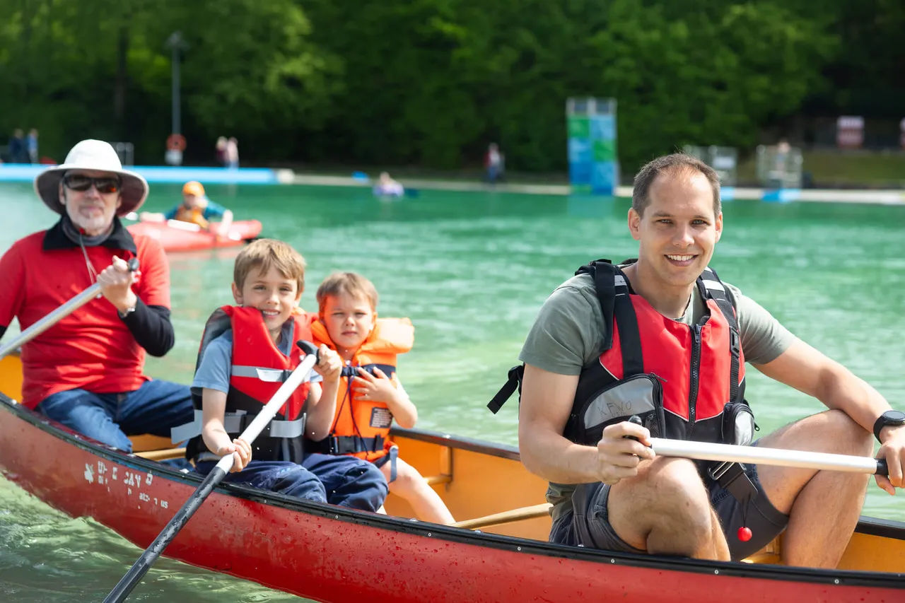 Vier Personen paddeln in einem Kanu auf dem See, der Mann am Steuer lächelt in die Kamera. Im Hintergrund weitere Boote und grüne Uferlandschaft.