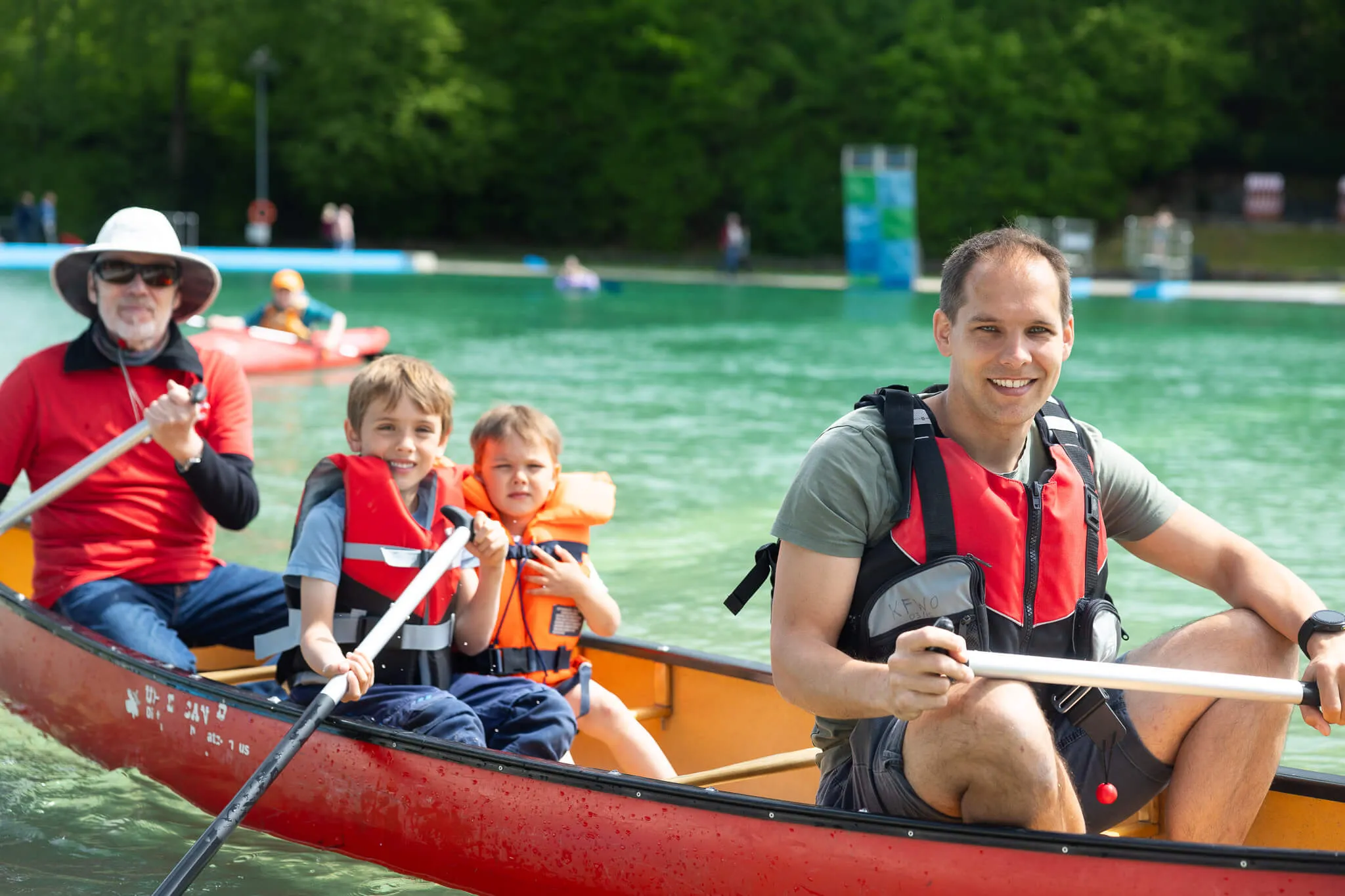 Vier Personen paddeln in einem Kanu auf dem See, der Mann am Steuer lächelt in die Kamera. Im Hintergrund weitere Boote und grüne Uferlandschaft.