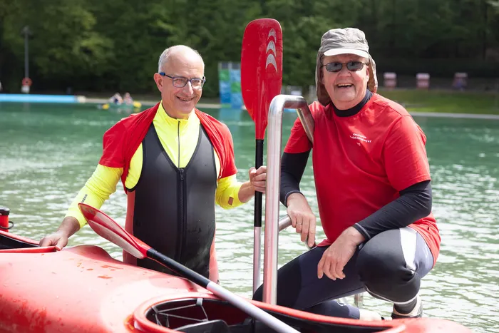 Zwei lächelnde Männer im Kajak halten Paddel, im Hintergrund weitere Boote auf dem Wasser und grüne Bäume.