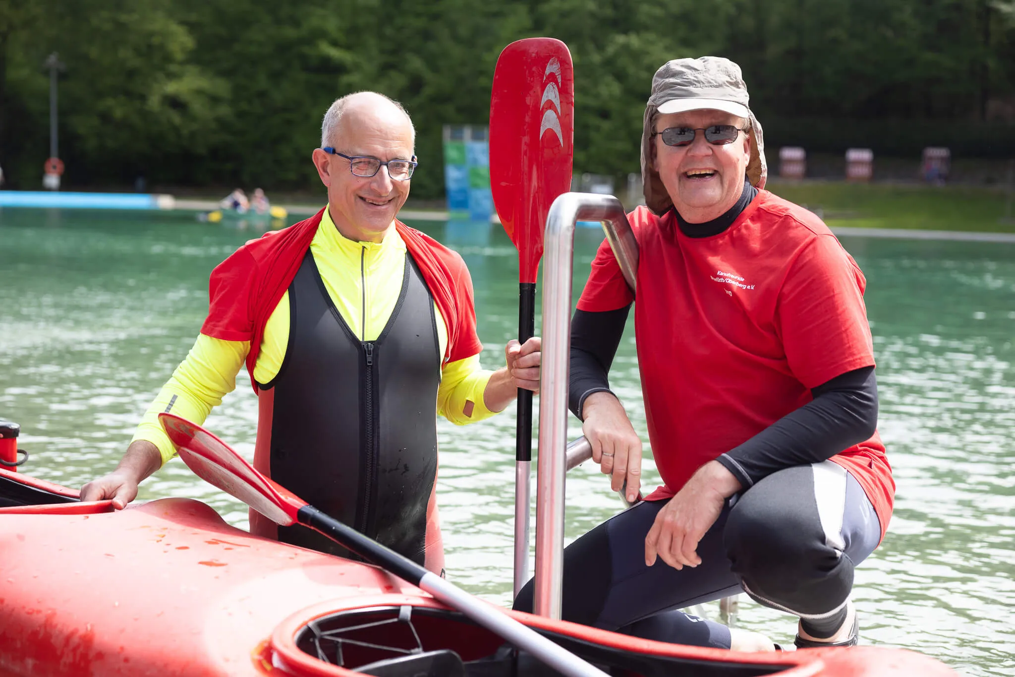 Zwei lächelnde Männer im Kajak halten Paddel, im Hintergrund weitere Boote auf dem Wasser und grüne Bäume.
