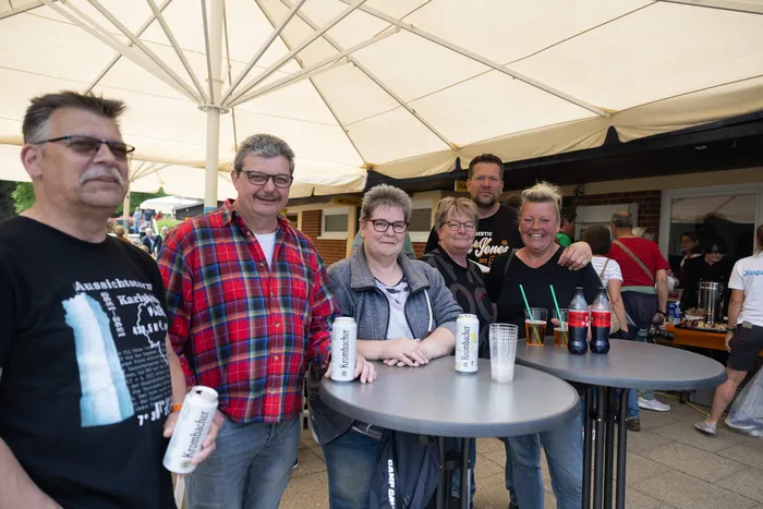 Eine Gruppe von Menschen steht lächelnd unter einem weißen Zelt und hält Bierflaschen. Im Hintergrund sind weitere Besucher und ein Gebäude zu sehen.