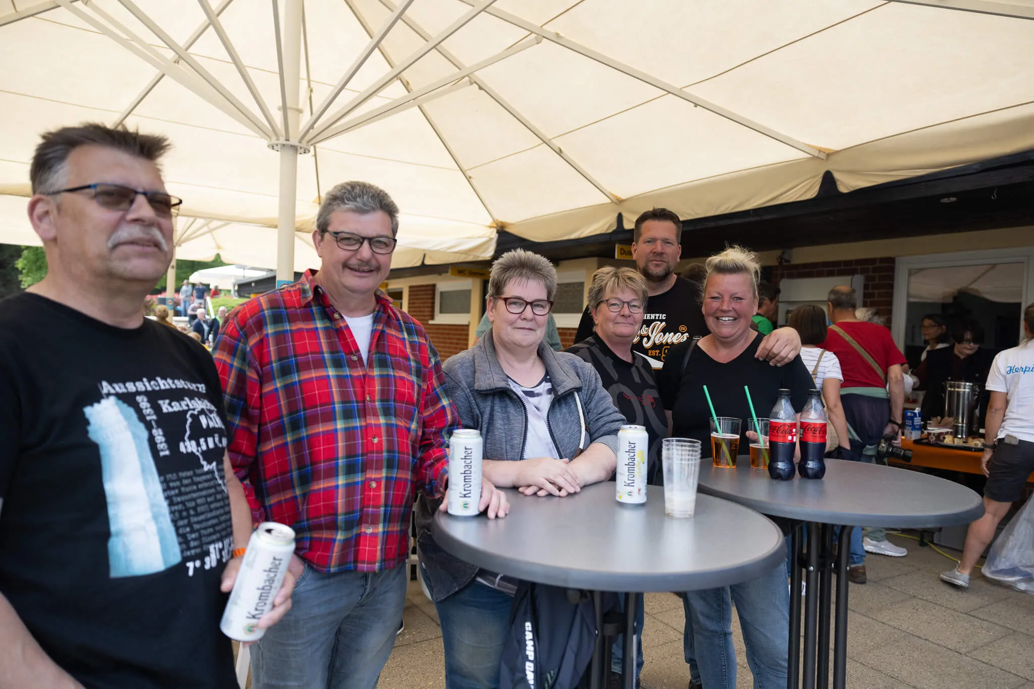 Eine Gruppe von Menschen steht lächelnd unter einem weißen Zelt und hält Bierflaschen. Im Hintergrund sind weitere Besucher und ein Gebäude zu sehen.