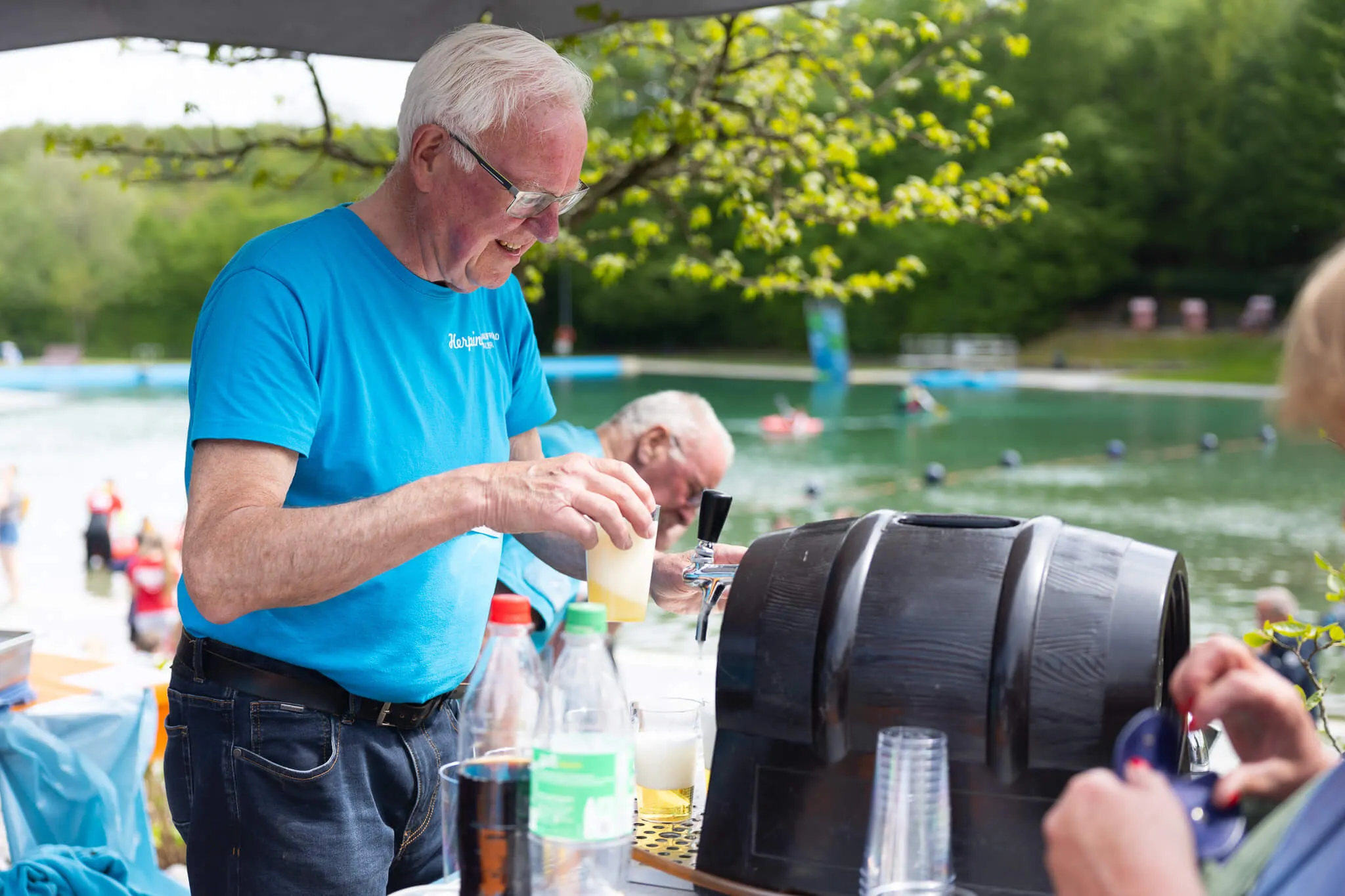 Ein älterer Mann mit blauem T-Shirt zapft Bier an einem Stehtisch, im Hintergrund baden Menschen in einem See.
