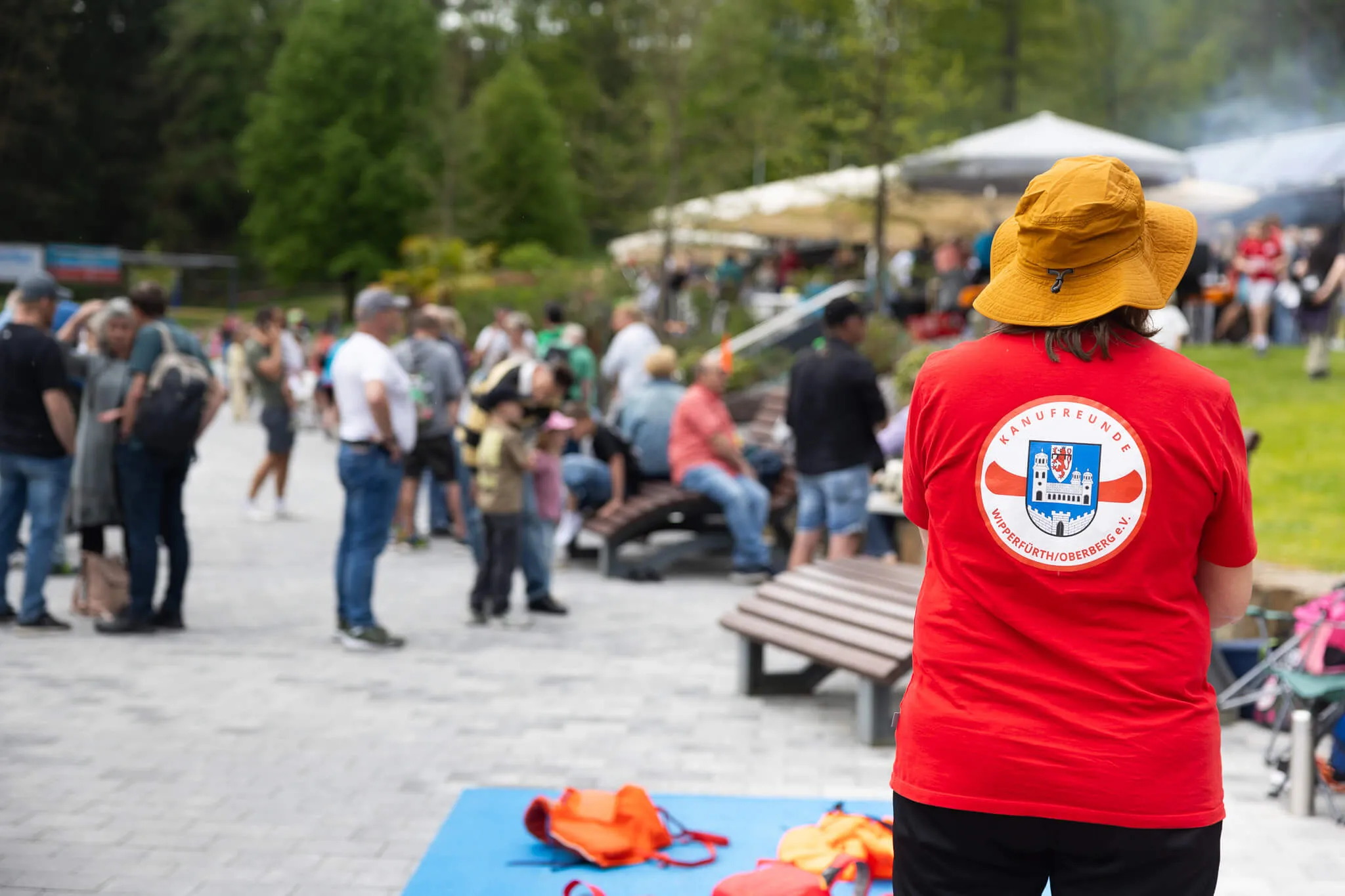 Rückansicht einer Person mit Hut und rotem T-Shirt mit dem Aufdruck „Kanufreunde Wipperfürth/Oberberg e.V.“ vor einer Menschenmenge auf einer Rasenfläche.