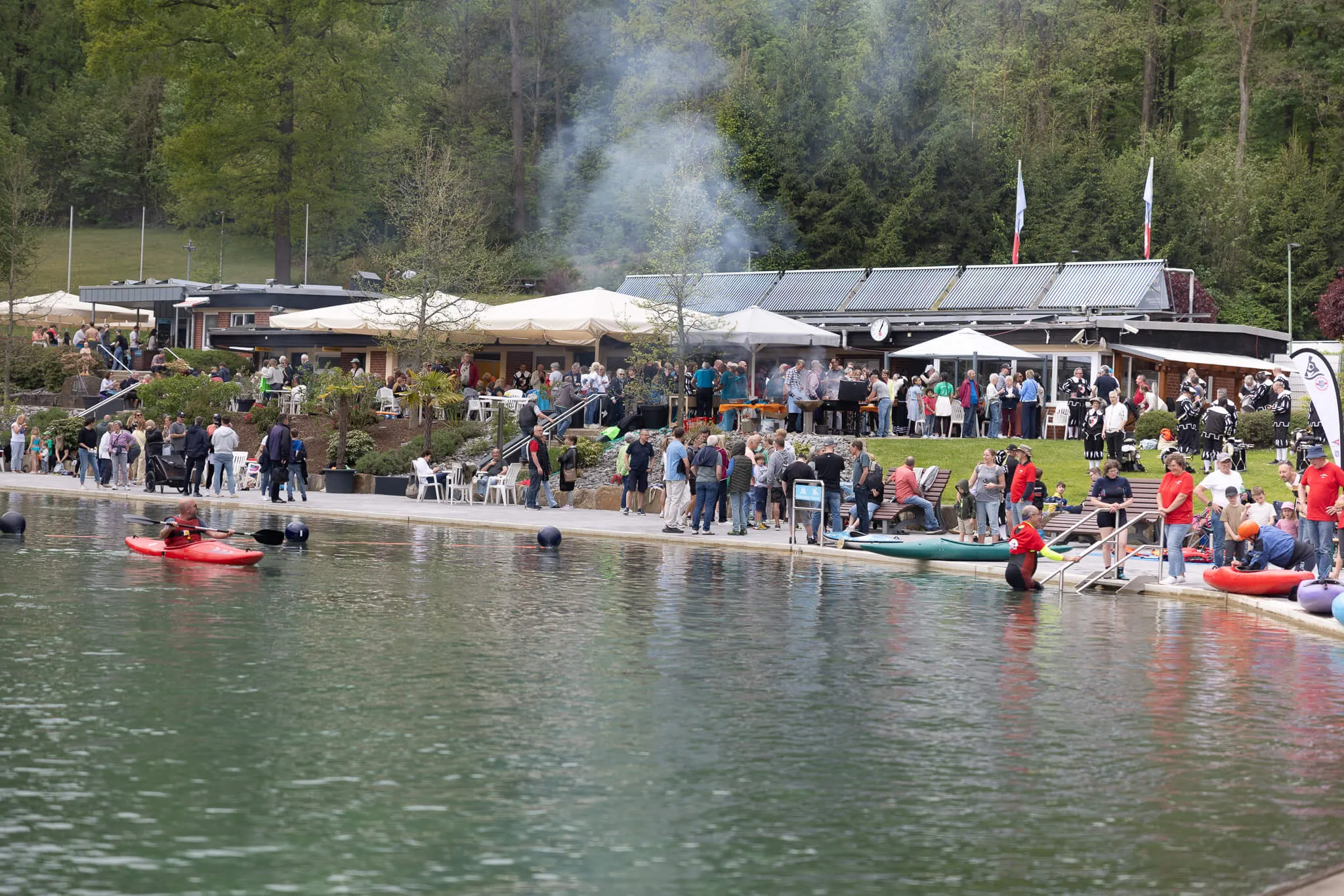 Blick auf die belebte Herpine in Halver zur Saisoneröffnung mit Besuchern an Tischen unter Sonnenschirmen und Booten auf dem See.
