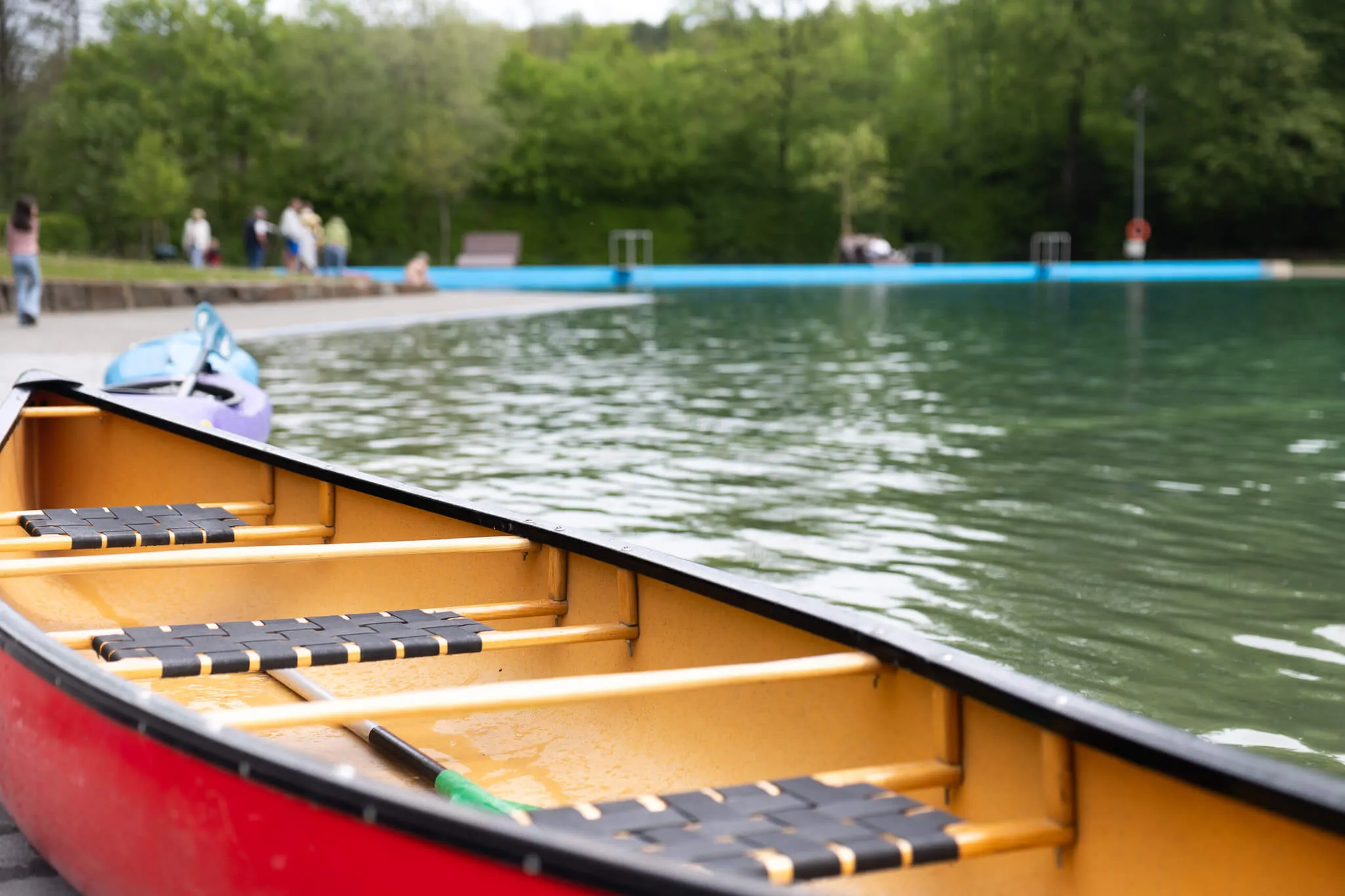 Rotes Kanu mit gelben Paddeln auf dem Wasser der Herpine in Halver, im Hintergrund Menschen und ein Freibad.