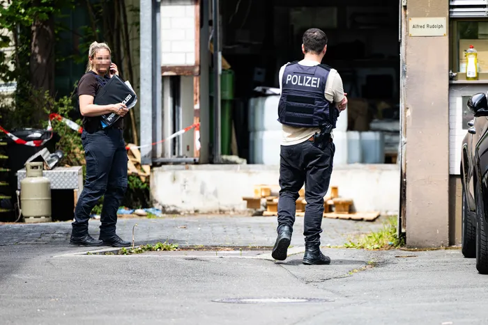Zwei Polizisten in Einsatzkleidung vor einem Gebäude mit Brandspuren und Absperrband. Im Hintergrund sind Chemikalienfässer zu sehen.