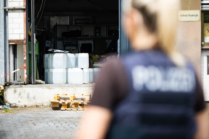 Ein Polizist steht mit dem Rücken zum Betrachter vor einer offenen Garagentür, hinter der mehrere weiße Kanister und Paletten mit Gegenständen zu sehen sind. Im Hintergrund ein Namensschild mit der Aufschrift „Alfred Rudolph“.