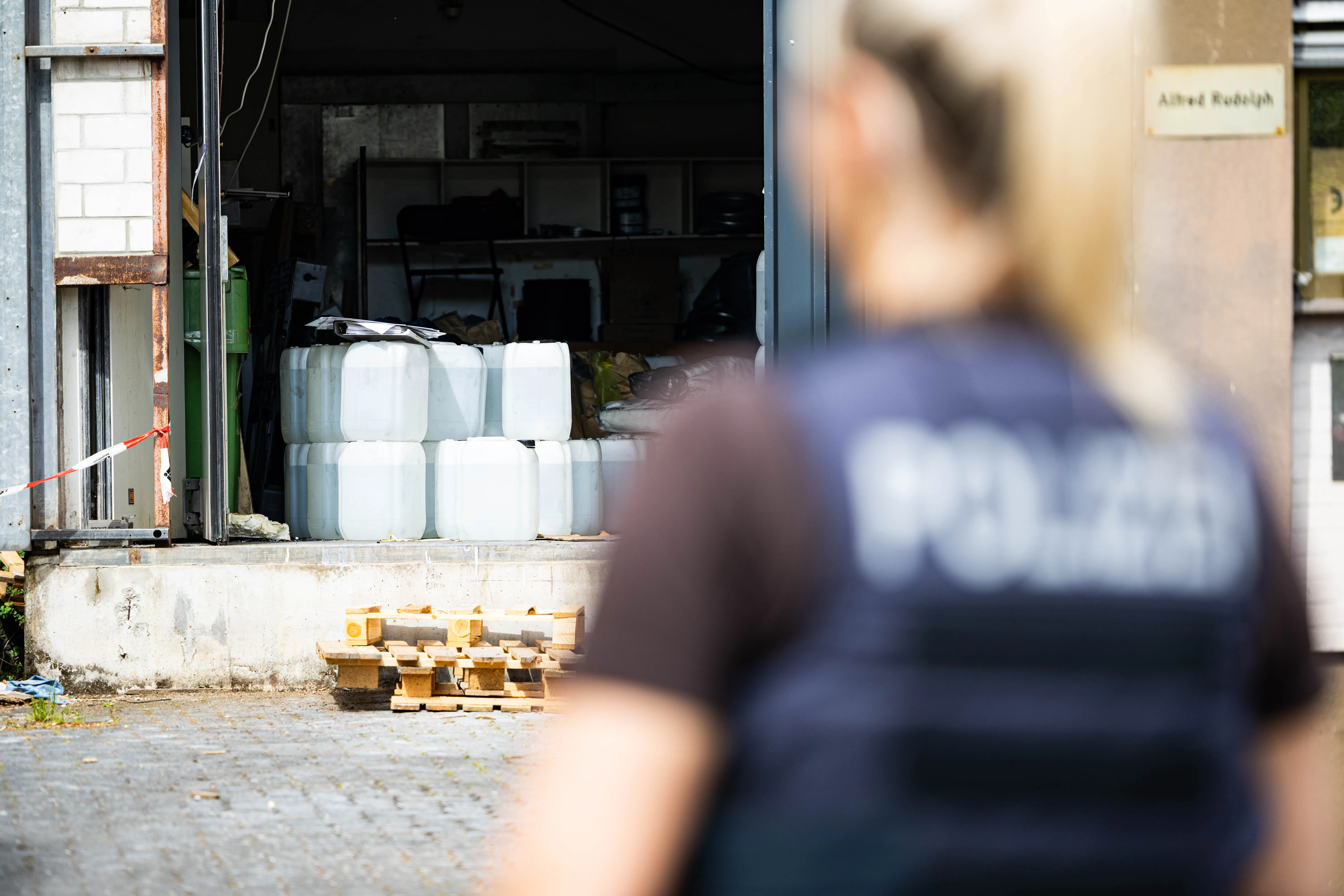Ein Polizist steht vor einer offenen Garagentür, hinter der mehrere weiße Pakete und Kanister zu sehen sind. Im Hintergrund ein Schild mit der Aufschrift „Alfred Rudolph“.