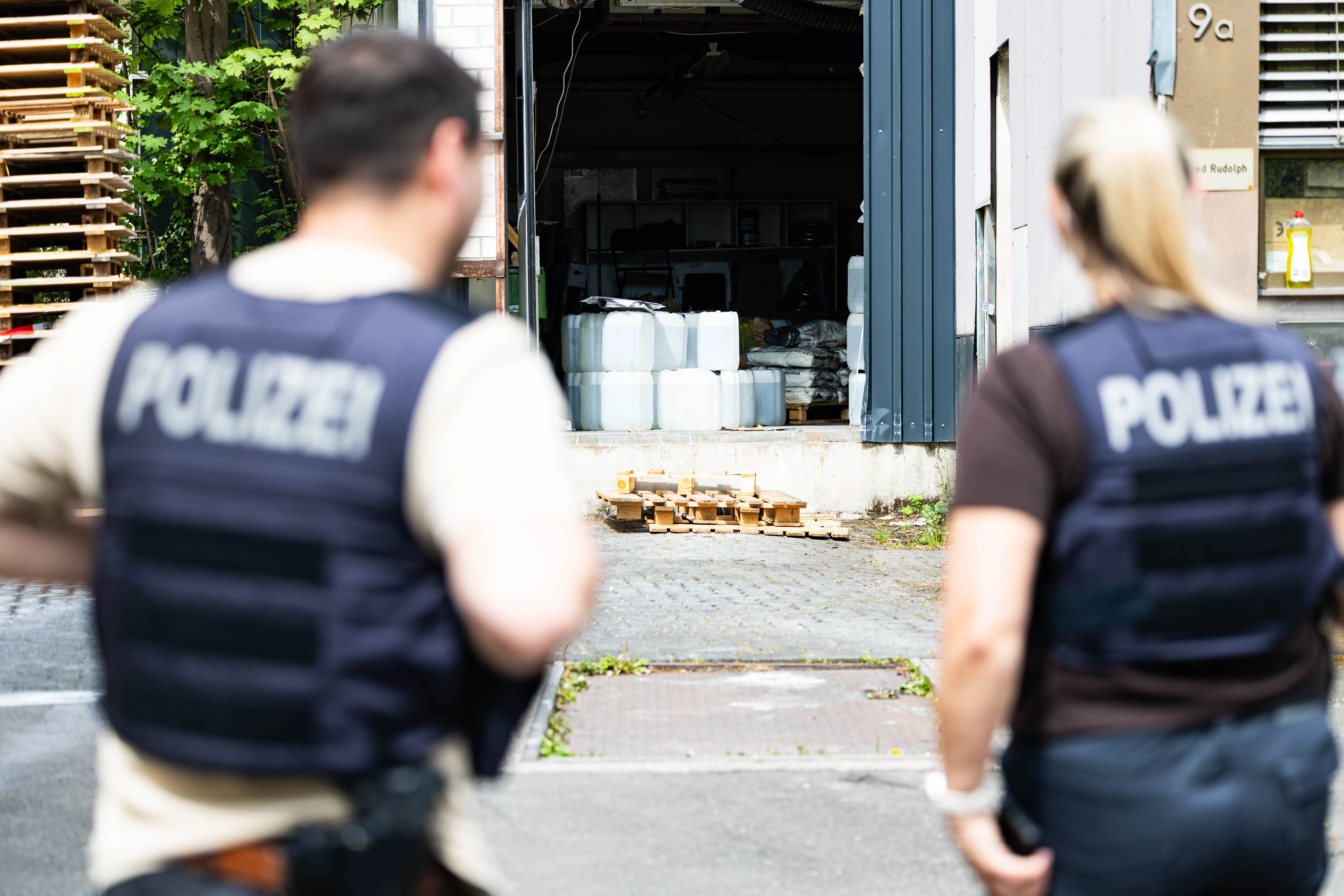 Zwei Polizisten mit beschrifteten Westen stehen vor einem offenen Lager mit gestapelten Chemikalienfässern.