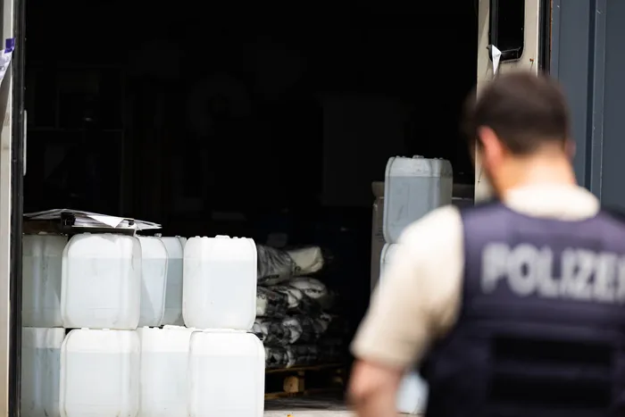 Polizist im Hintergrund vor gestapelten weißen Kanistern und weiteren Chemikalien in einem Lagerraum, möglicherweise Spuren eines illegalen Drogenlabors.