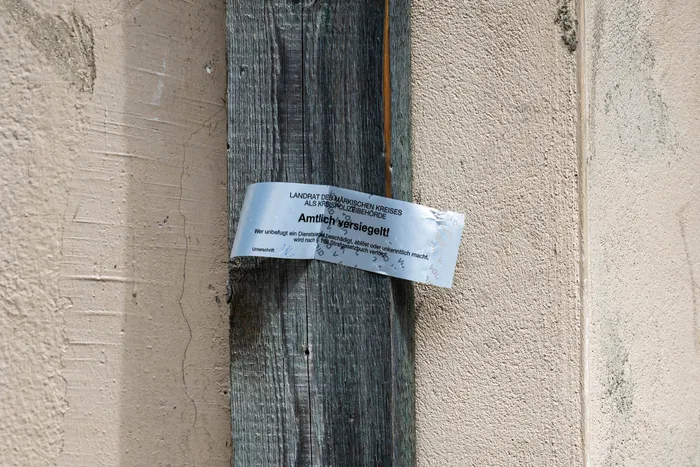 Ein weißes, mit Warnhinweisen versehenes Siegel ist an einem Holzmast befestigt. Der Text auf dem Siegel warnt davor, die Versiegelung zu beschädigen oder zu entfernen, da dies strafbar ist.