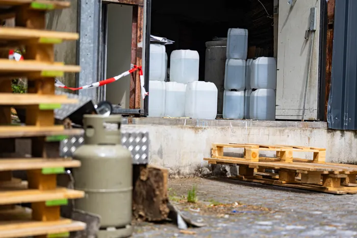 Vor einer offenen Lagerhalle stehen mehrere Kanister und eine Gasflasche auf dem Hof. Eine Absperrung mit rotem Band ist zu sehen.