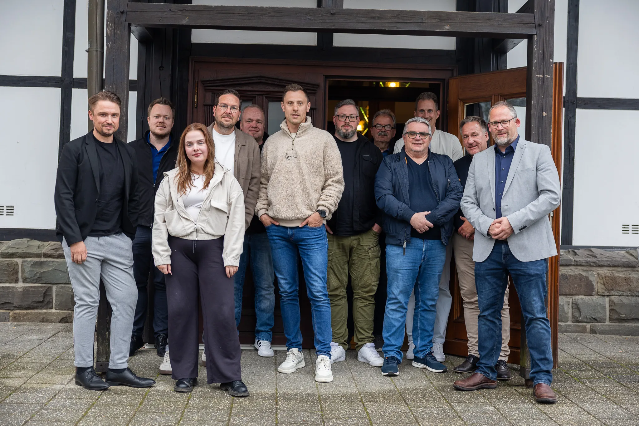 Gruppe von zehn Personen steht vor einem Fachwerkhauseingang zusammen. Sie sind unterschiedlich gekleidet, einige tragen Anzüge, andere Jeans und Pullover.