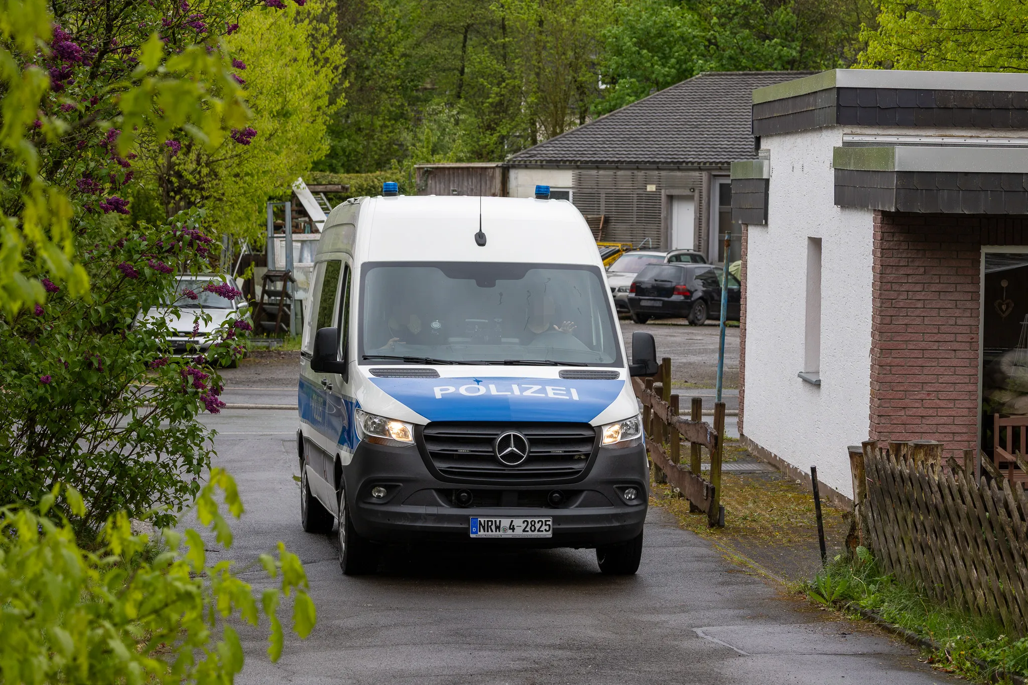 Weißer Polizei-Transporter steht auf nasser Straße vor einem Gebäude, im Hintergrund weitere Fahrzeuge und Bäume mit blühenden Zweigen.