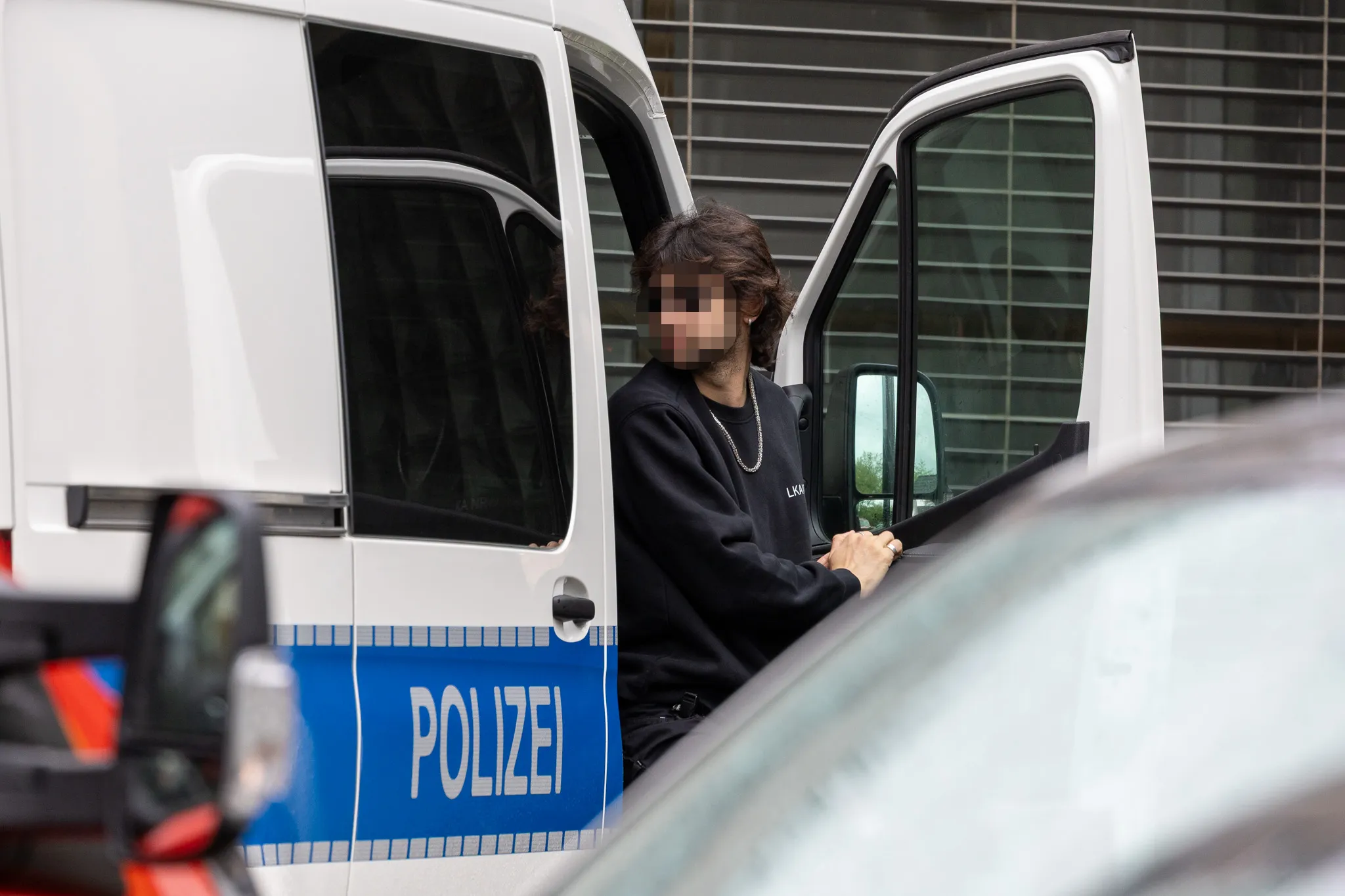 Eine Person mit verpixelter Gesichtsansicht steigt aus einem geöffneten Transporter der Polizei. Im Hintergrund ein Gebäude mit Glasfassade.
