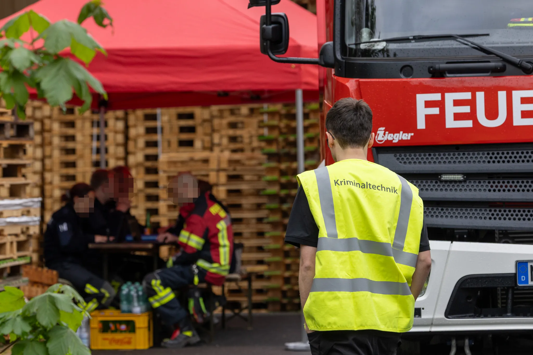 Ein Mitarbeiter des Landeskriminalamts mit Warnweste steht vor einem roten Feuerwehrauto, während im Hintergrund unter einem Zelt Polizisten arbeiten.