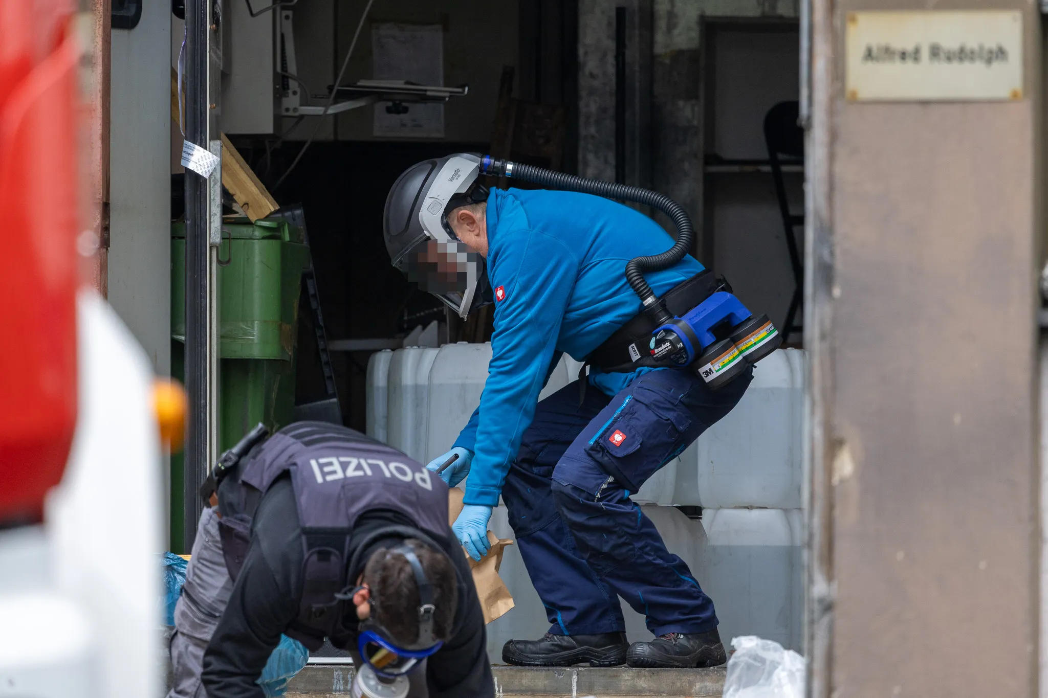 Kriminaltechniker in Schutzanzügen untersuchen einen Raum, vermutlich ein Drogenlabor, mit Geräten und Müllcontainern.