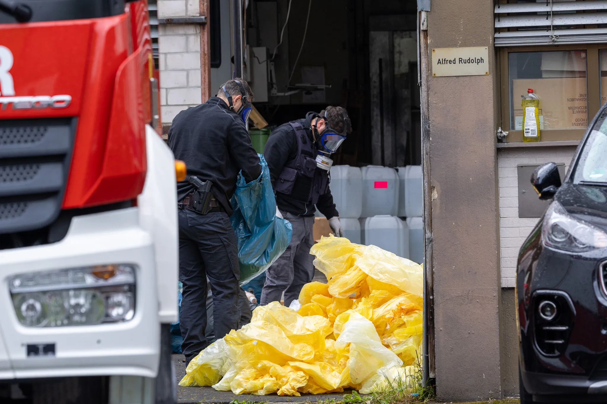 Polizisten in Schutzanzügen untersuchen vor einem Gebäude und einem roten Lkw eine Ansammlung gelber Folien und Behälter.