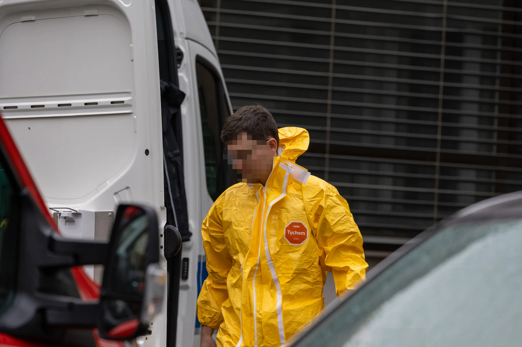 Person in gelbem Schutzanzug mit dem Aufdruck „Tychem“ vor einem weißen Transporter und einem Auto.