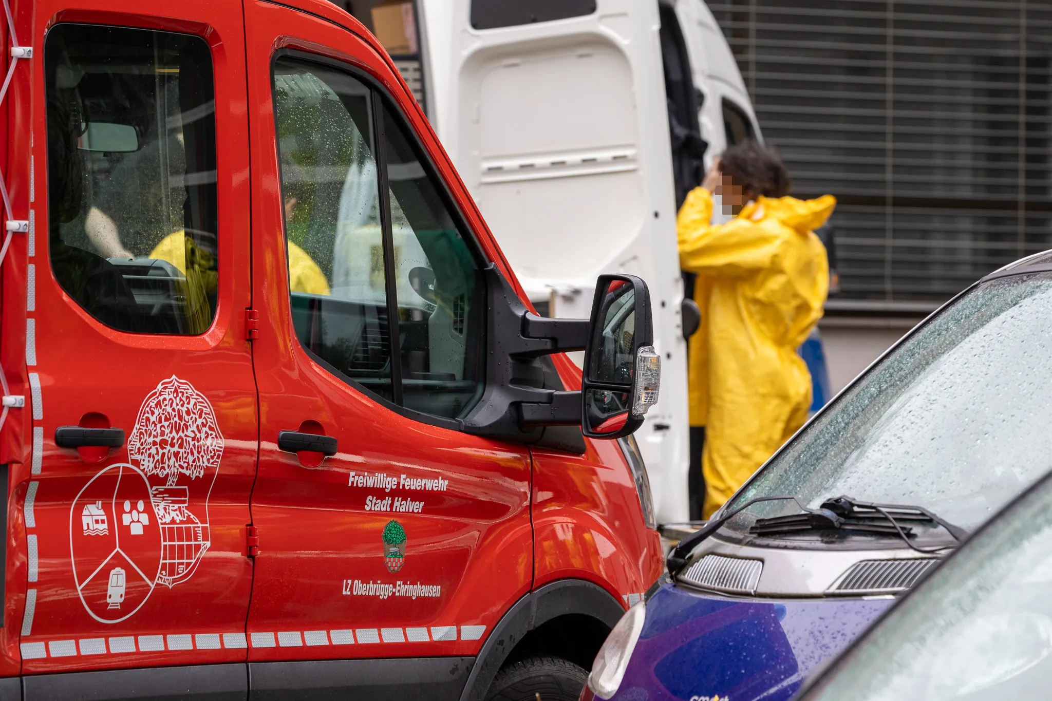 Rotes Fahrzeug der Freiwilligen Feuerwehr Stadt Halver steht neben einem blauen Auto. Im Hintergrund sind Personen in Schutzanzügen bei der Arbeit zu sehen.