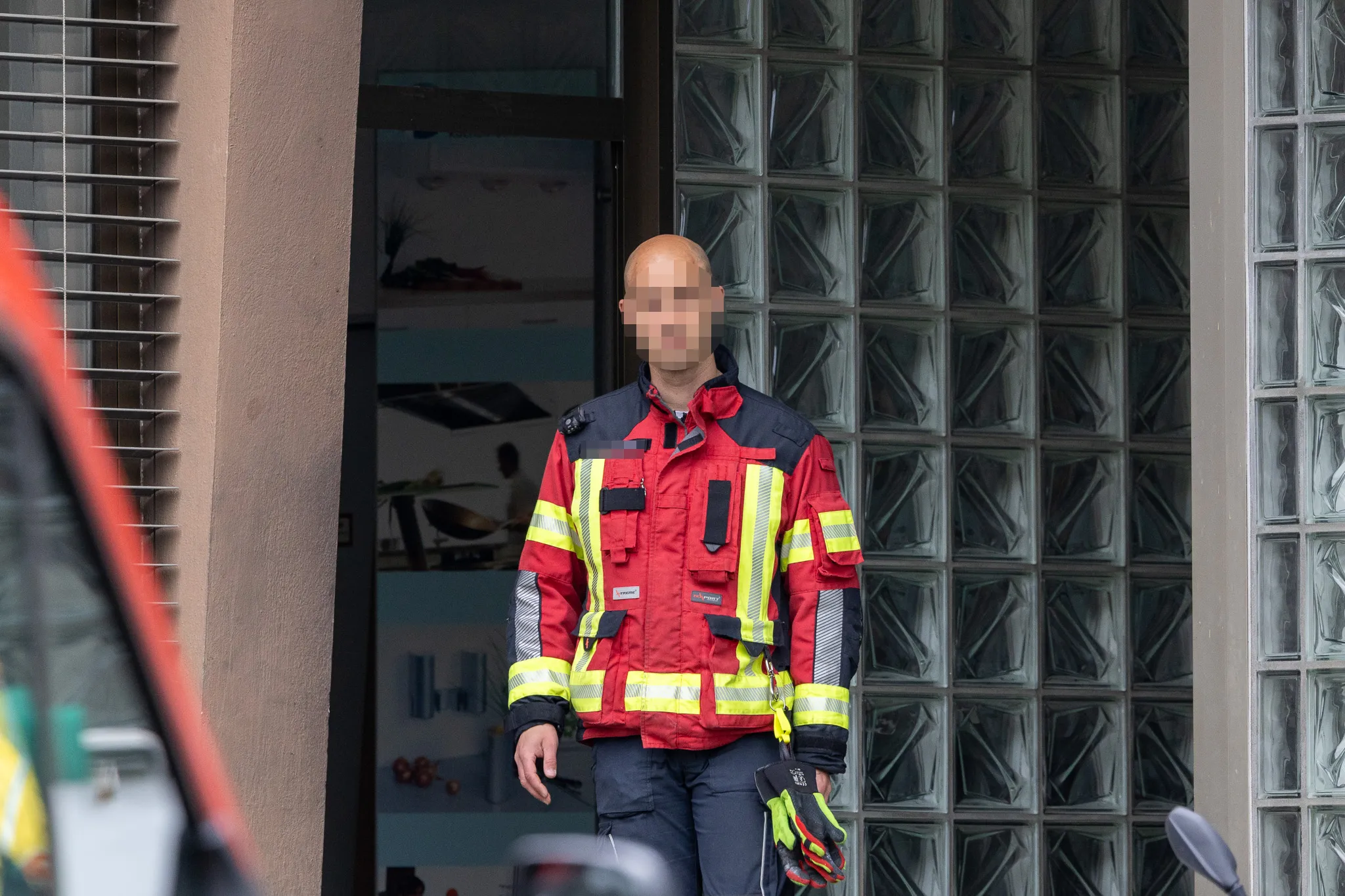 Feuerwehrmann in roter Einsatzjacke mit gelben Reflexstreifen steht vor einem Gebäude mit Glasbausteinen.