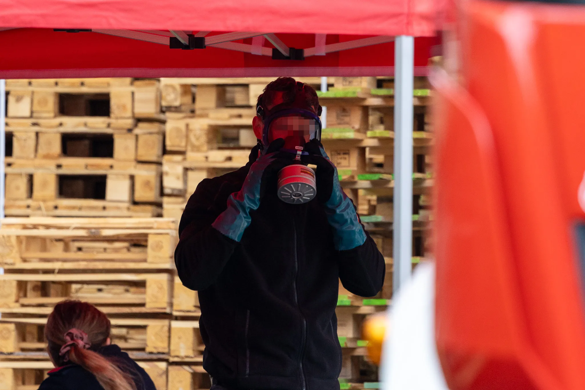 Eine Person mit Schutzbrille und Atemschutzmaske hält ein rot-weißes Messgerät vor sich. Im Hintergrund sind Holzpaletten und ein rotes Zeltdach zu sehen.