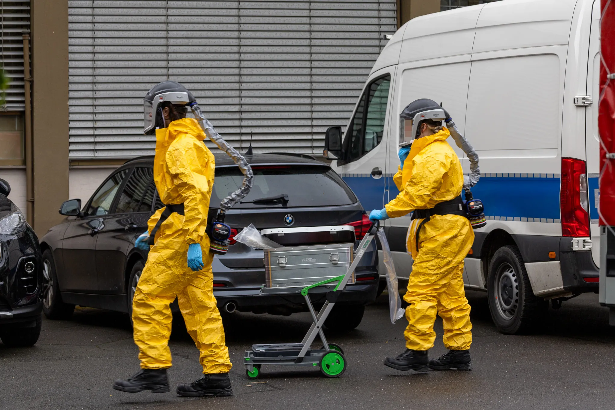 Zwei Personen in gelben Schutzanzügen mit Atemschutzmasken stehen vor einem geöffneten Kofferraum eines BMW und einem Transporter.