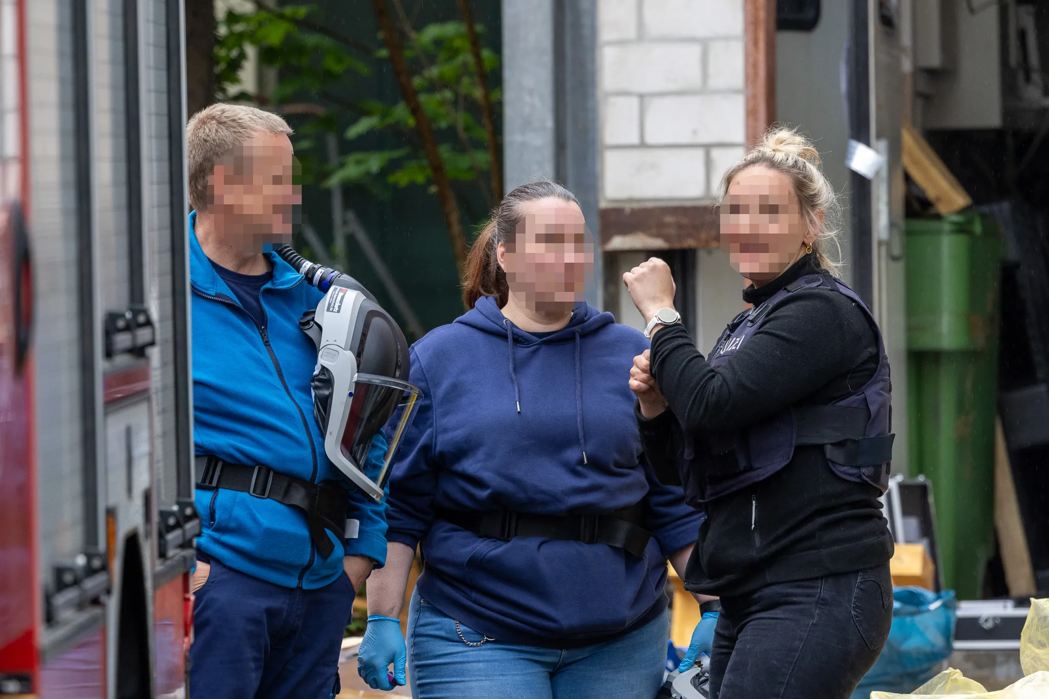 Zwei Polizistinnen in Schutzwesten führen eine Frau mit hochgebundenen Haaren ab, während ein Sanitäter im Hintergrund steht. Im Hintergrund sind ein Einsatzwagen und Mülltonnen zu sehen.