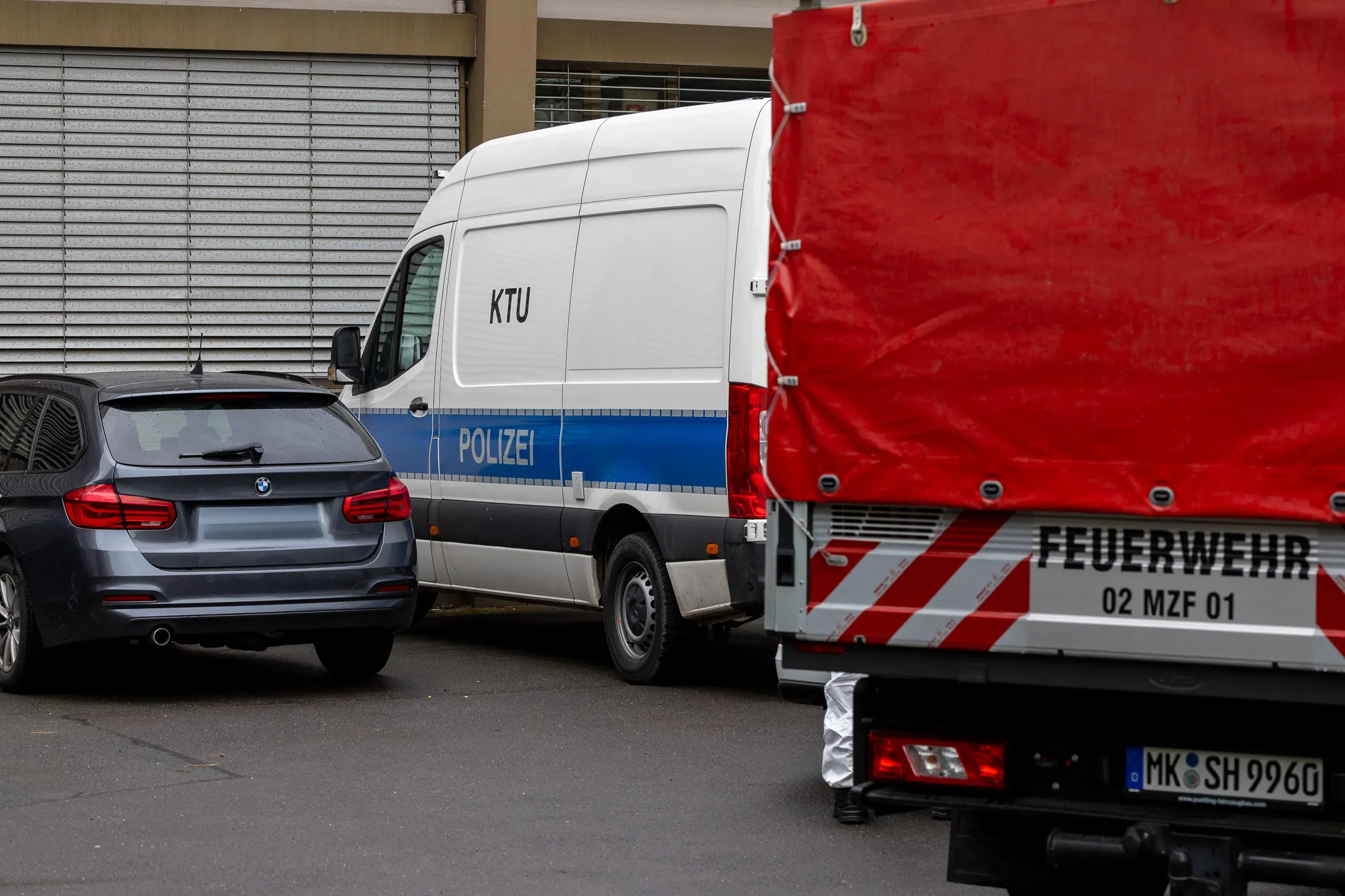 Polizeivan und Fahrzeug der Feuerwehr stehen auf einem Parkplatz vor einem Gebäude mit geschlossenen Rollläden. Im Hintergrund ist ein schwarzer BMW zu sehen.
